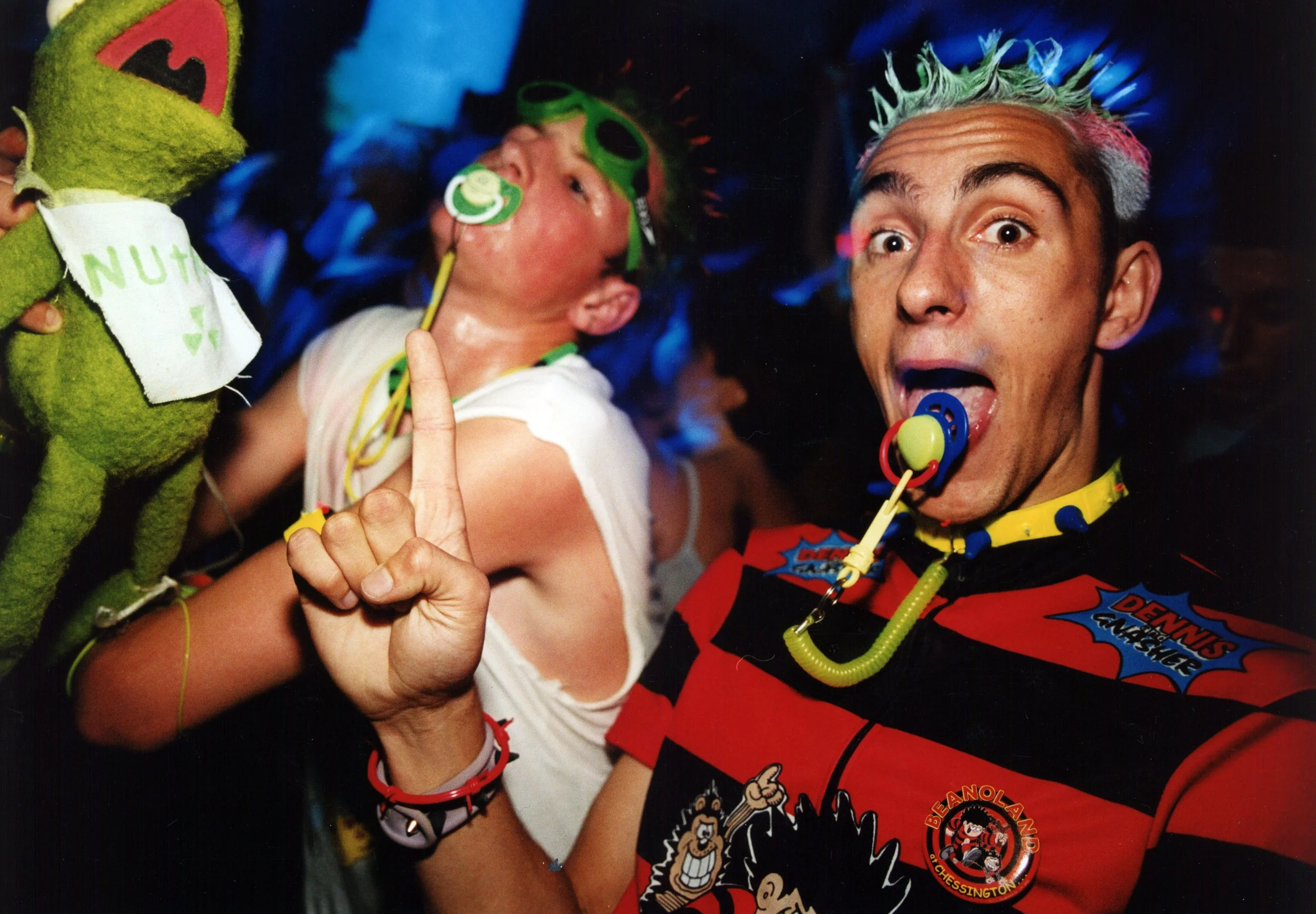 A young boy with a pacifier and goggles, holding a hand up with one finger, next to an adult man with colorful hair and a pacifier, both dressed in colorful costumes at a lively event or party.