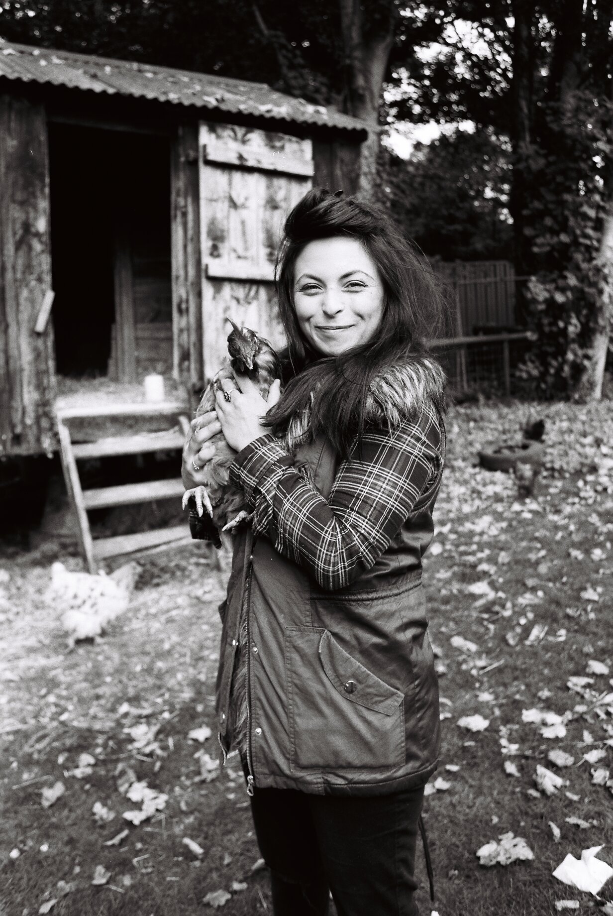 A woman with long hair smiling and holding a chicken outdoors near a wooden chicken coop with trees in the background.