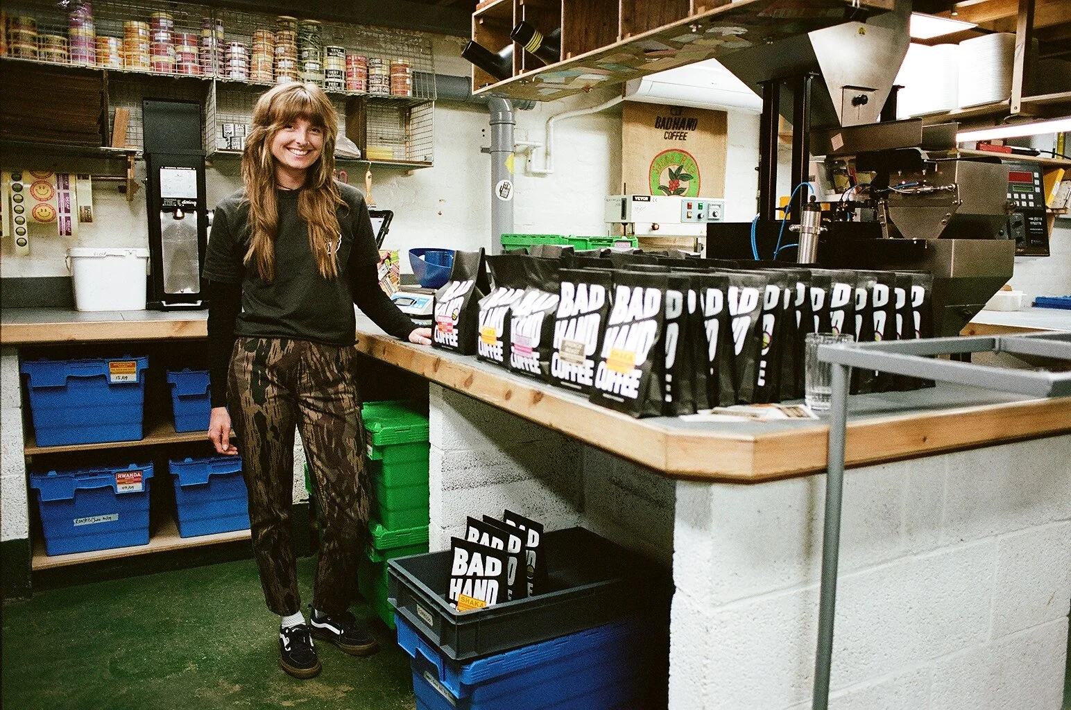 Bad Hand Coffee team member standing beside branded coffee bags and roasting equipment in the production workspace. Photo by Shahab Notghi.