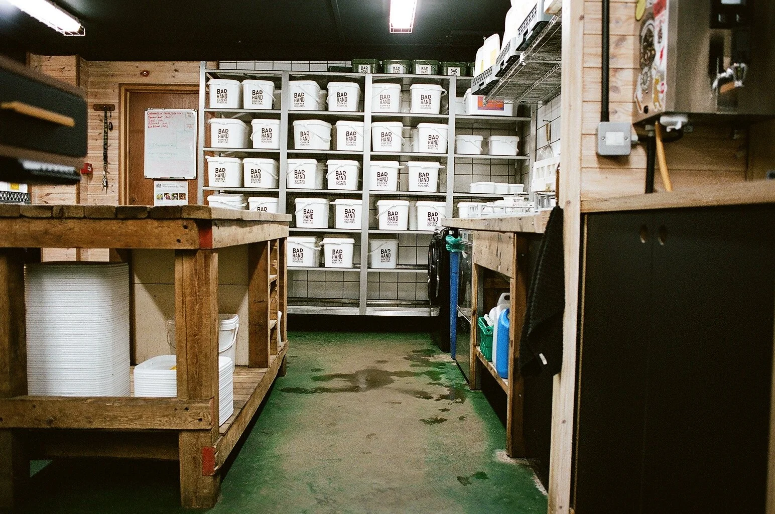 Storage area inside Bad Hand Coffee with shelves of branded white coffee containers and wooden workbenches. Photo by Shahab Notghi.