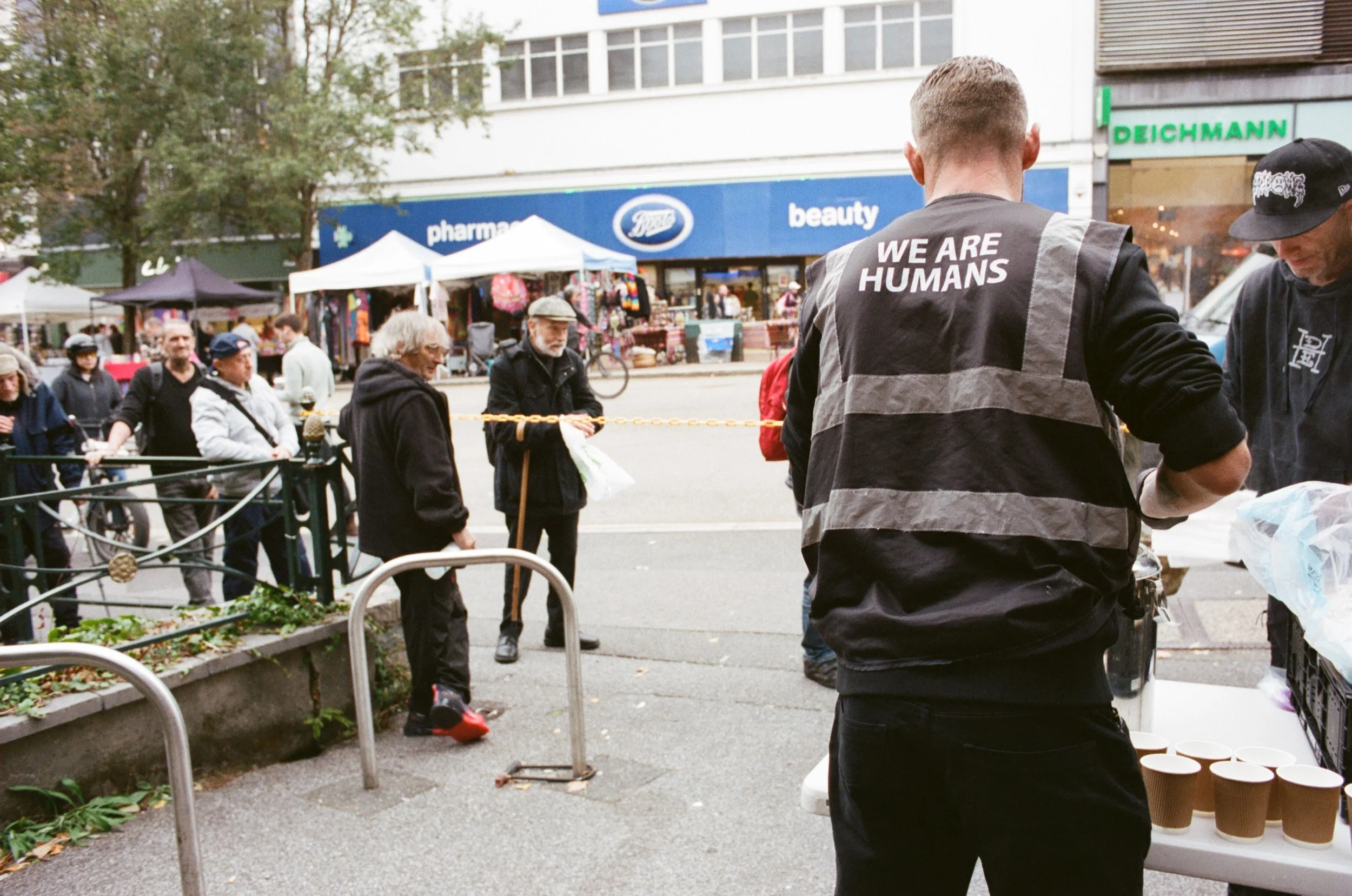 People waiting in line outside a market, with tents and a sign for a pharmacy in the background. An individual with a jacket reading 'WE ARE HUMANS' is in the foreground.