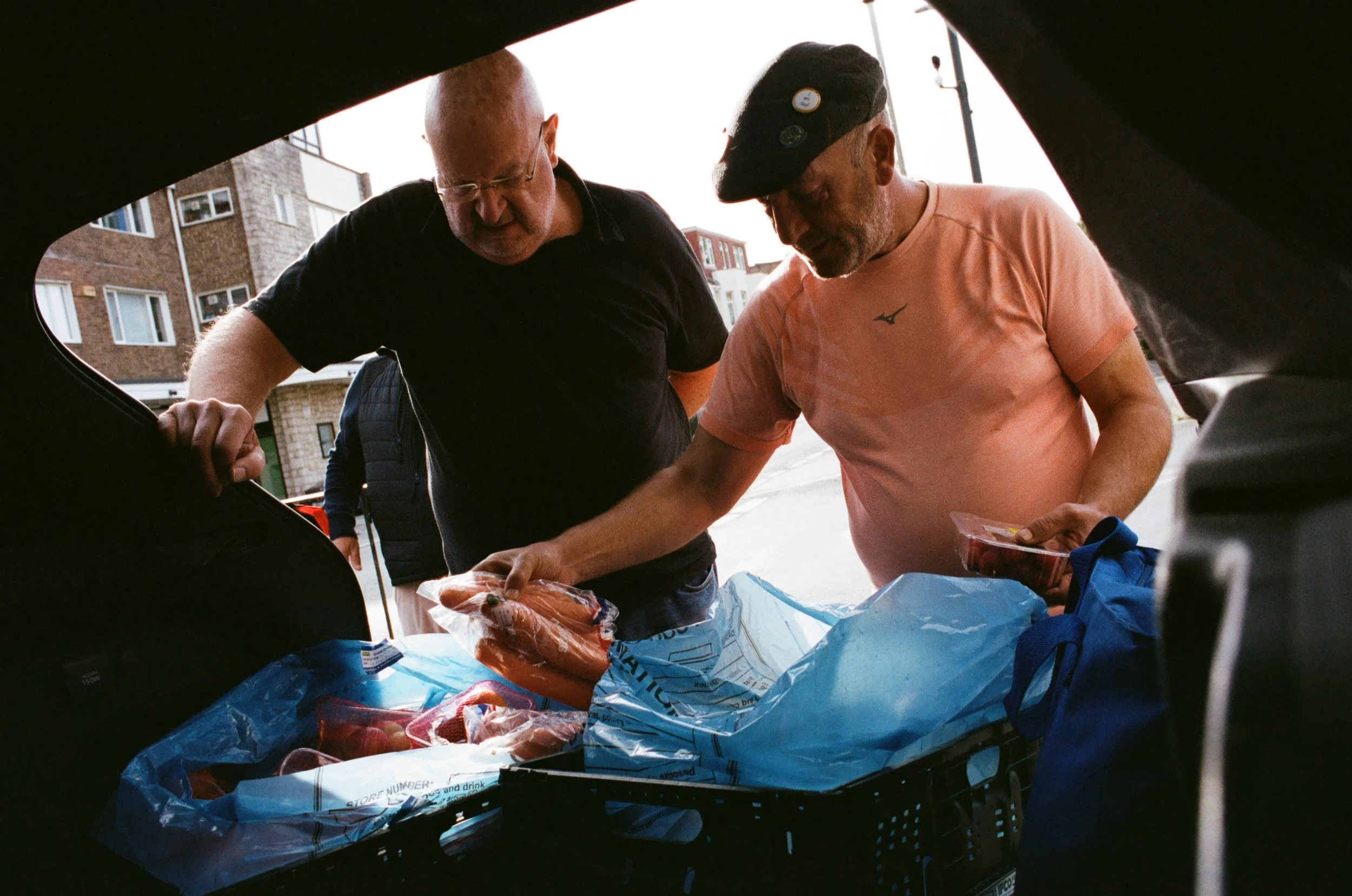 Two men are leaning over the open trunk of a vehicle, packing or unpacking grocery bags containing food items. The scene is outdoors with apartment buildings in the background.