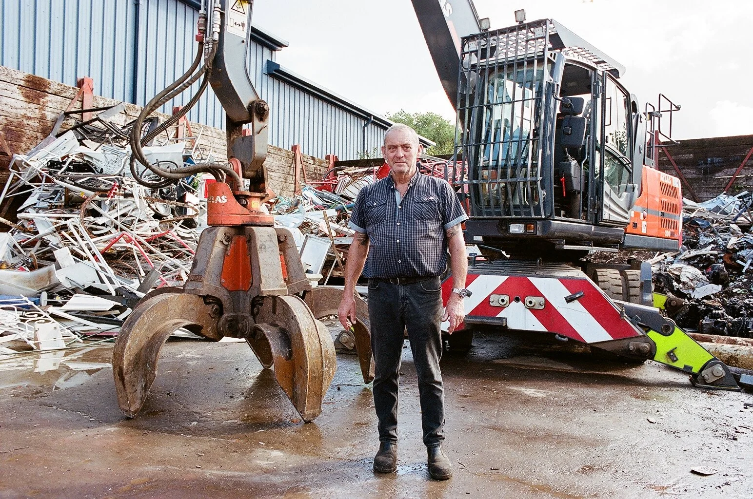 A man standing in front of a large orange and black industrial machine at a scrapyard, with a pile of scrap metal behind him.