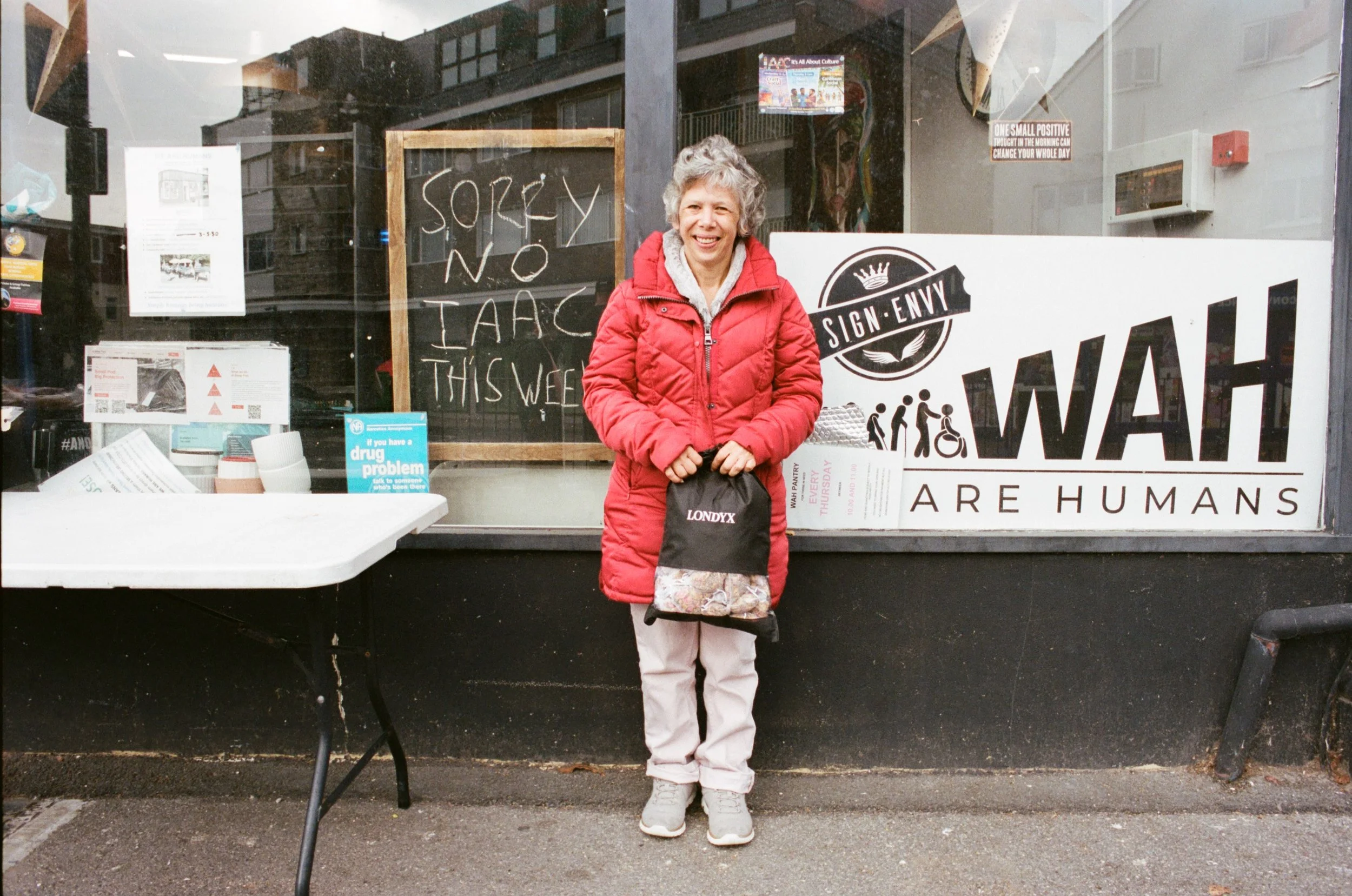 A smiling woman with gray hair wearing a red jacket and light-colored pants, standing in front of a storefront window with signs, including a chalkboard saying 'SORRY NO TAAC THIS WEEK,' and a large logo that says 'SIGN ENNVY' with the slogan 'WHAT A