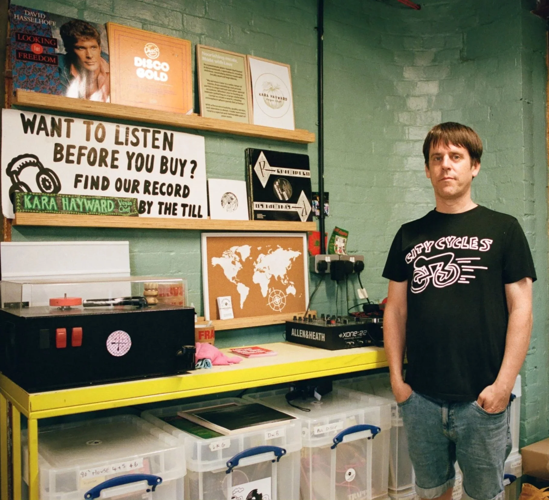 Man standing beside record displays and colourful signage inside a shop with green walls. Photo by Shahab Notghi.