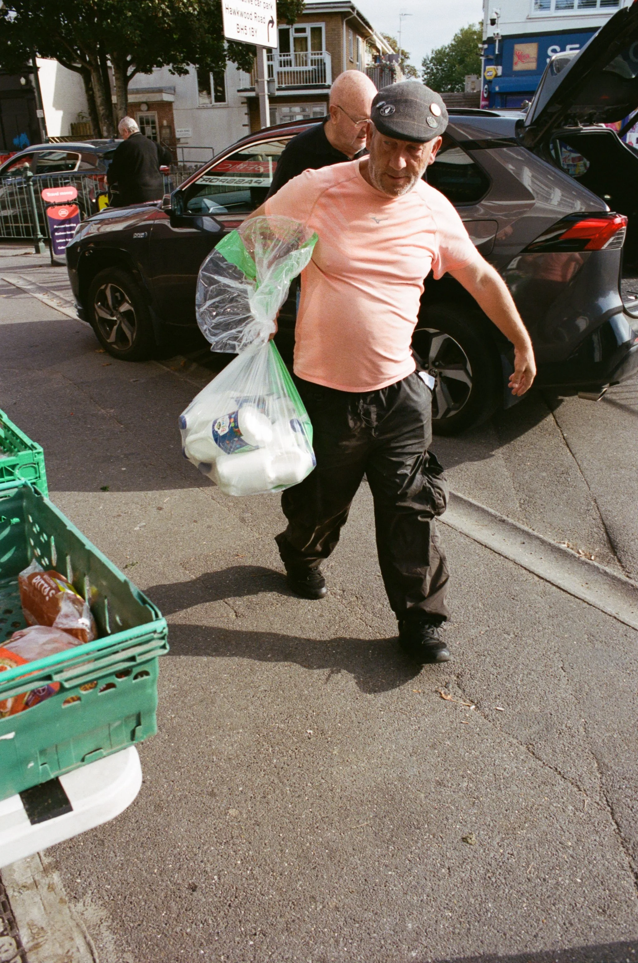 A man with a gray cap and peach T-shirt carrying a bag of groceries walks on a city sidewalk, with a parked black car and other pedestrians in the background.