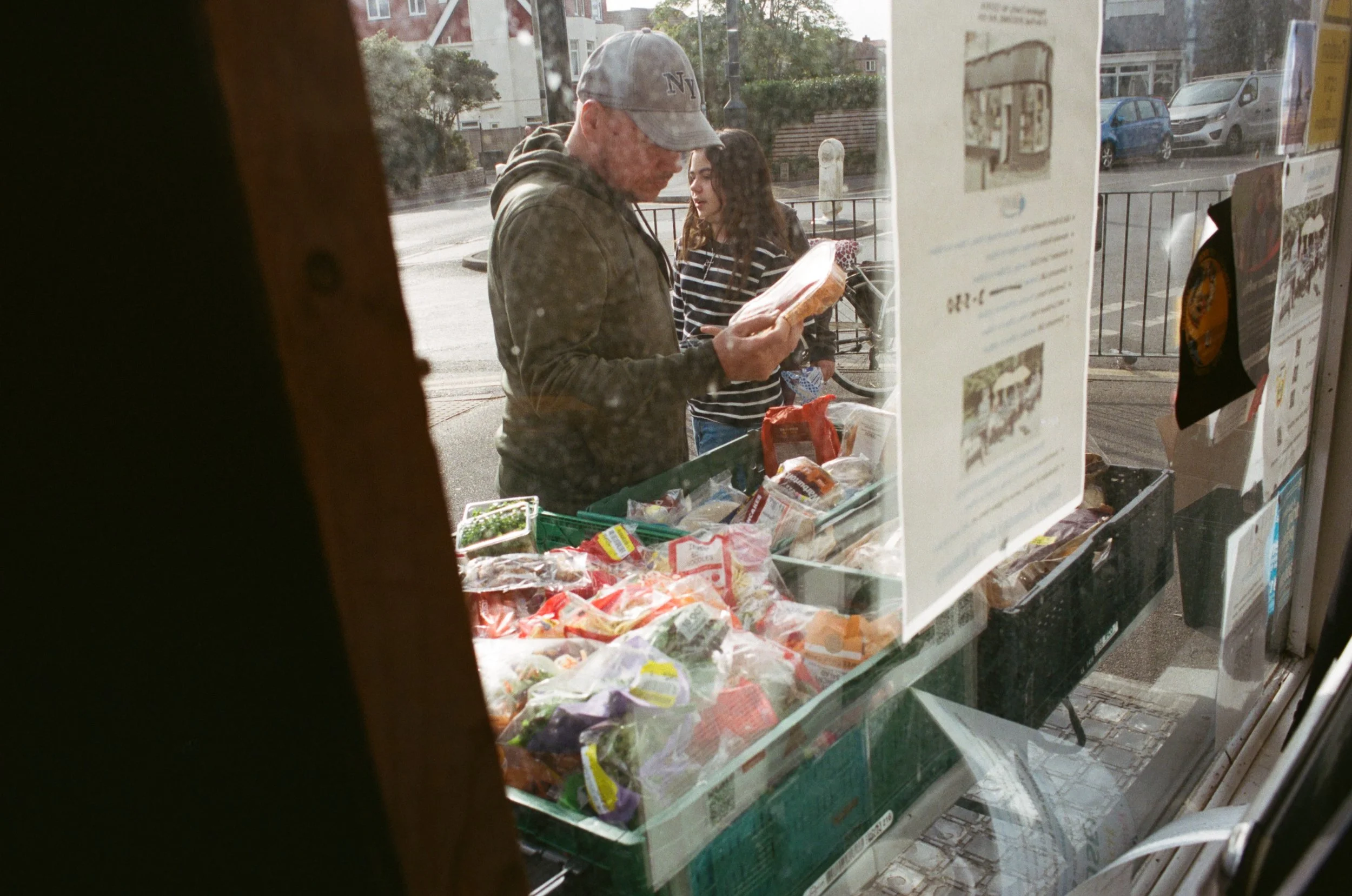 A man wearing a gray baseball cap and a gray hoodie is inside a store, looking at food items. A young woman with long brown hair, wearing a striped shirt, stands behind him. Outside the store window, parked cars and buildings are visible.