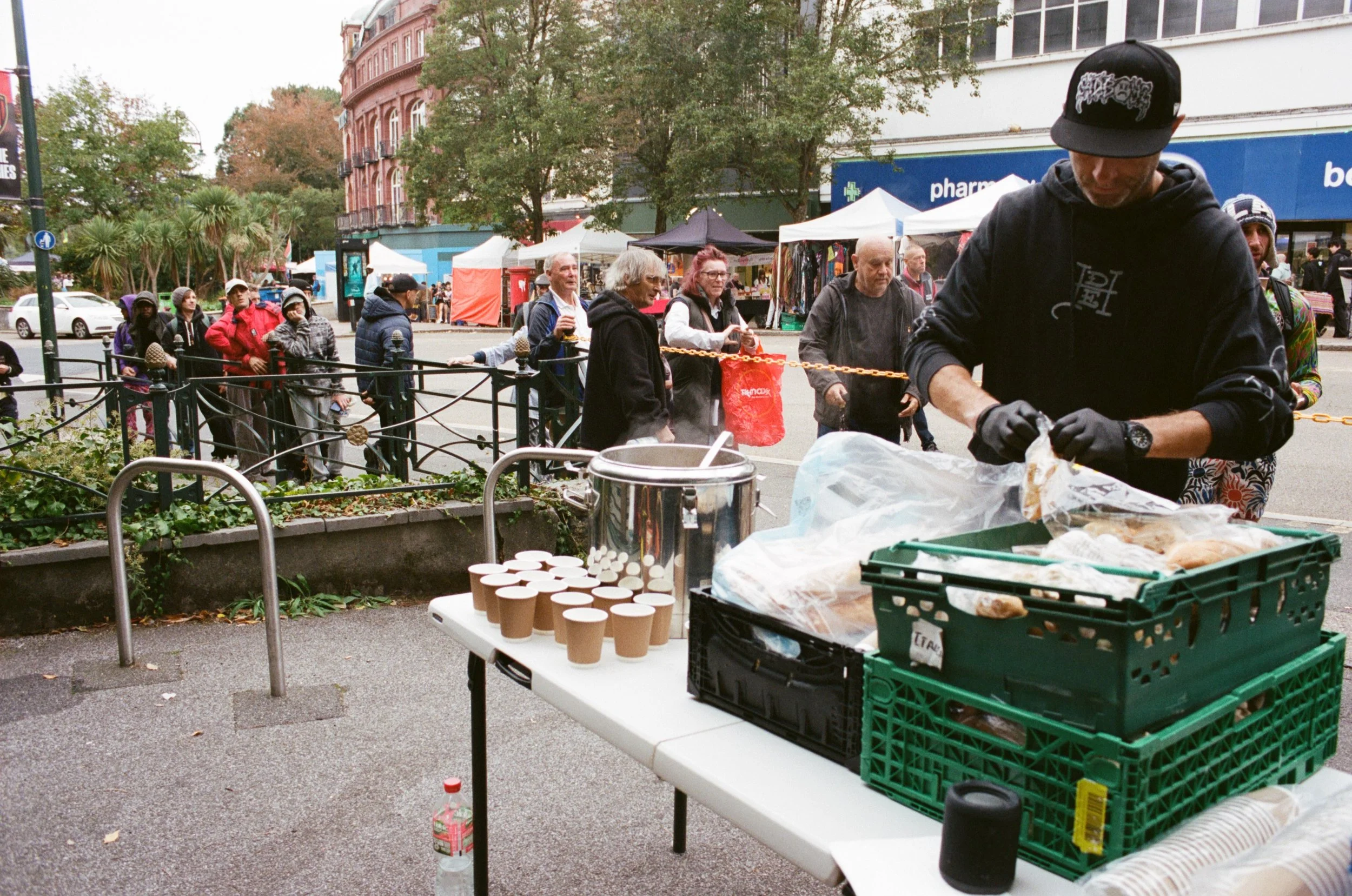 A street scene with people waiting in line at an outdoor market. There are tents and stalls set up selling various goods, and a table with cups and containers. The background includes trees, buildings, and cars.