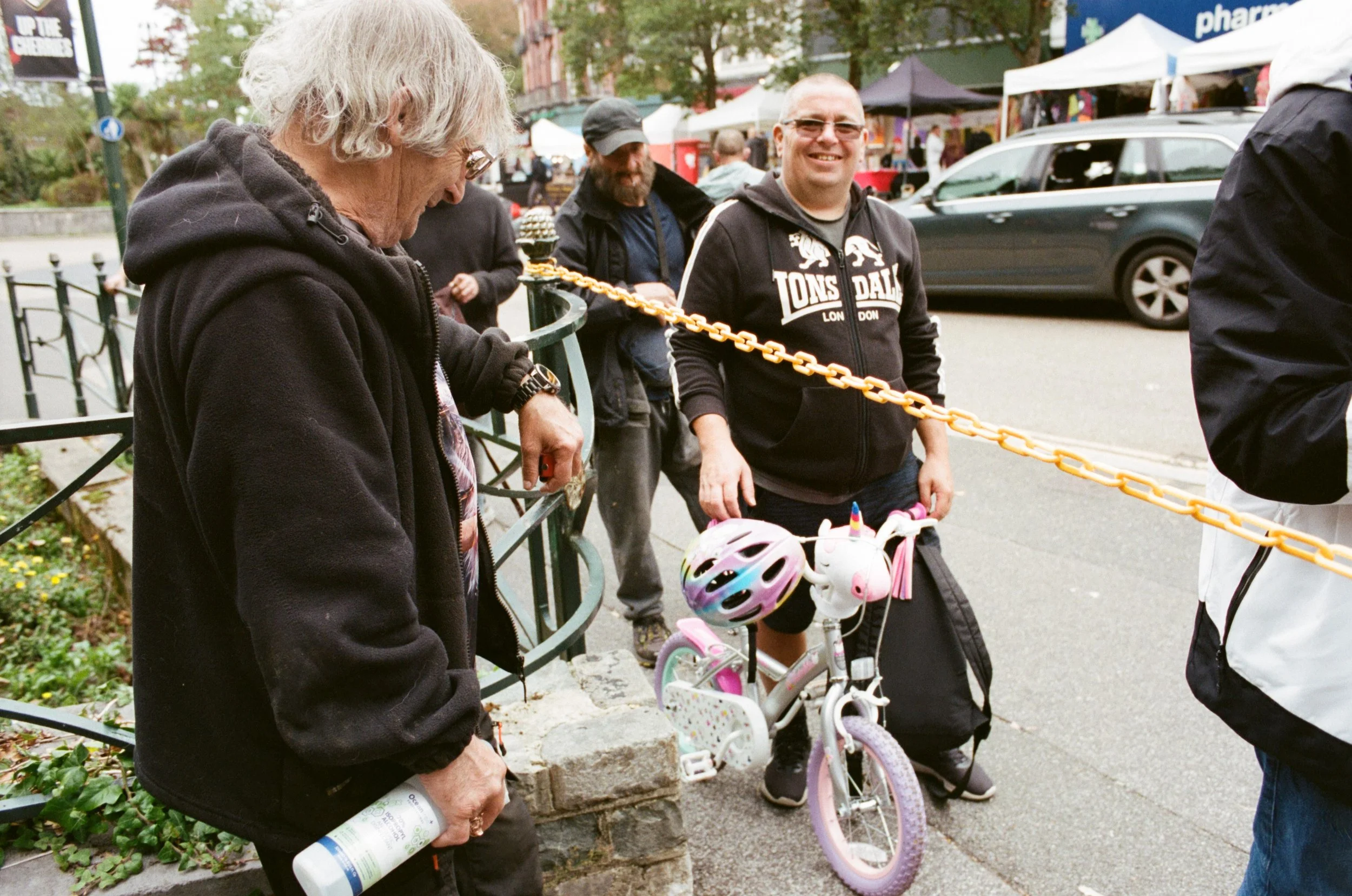 A group of people standing on a street, with tents and cars in the background, including a young girl with a pink bike and helmets, and three adult men, one with curly gray hair holding a water bottle.