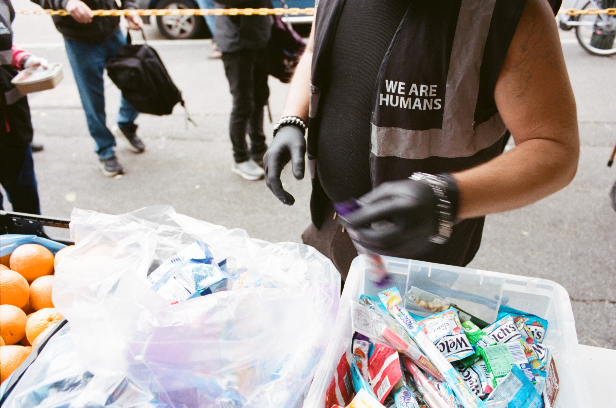 Close-up of a person wearing a vest with a message 'We Are Humans' at an outdoor market or event, with people in the background and various snack items on a table.