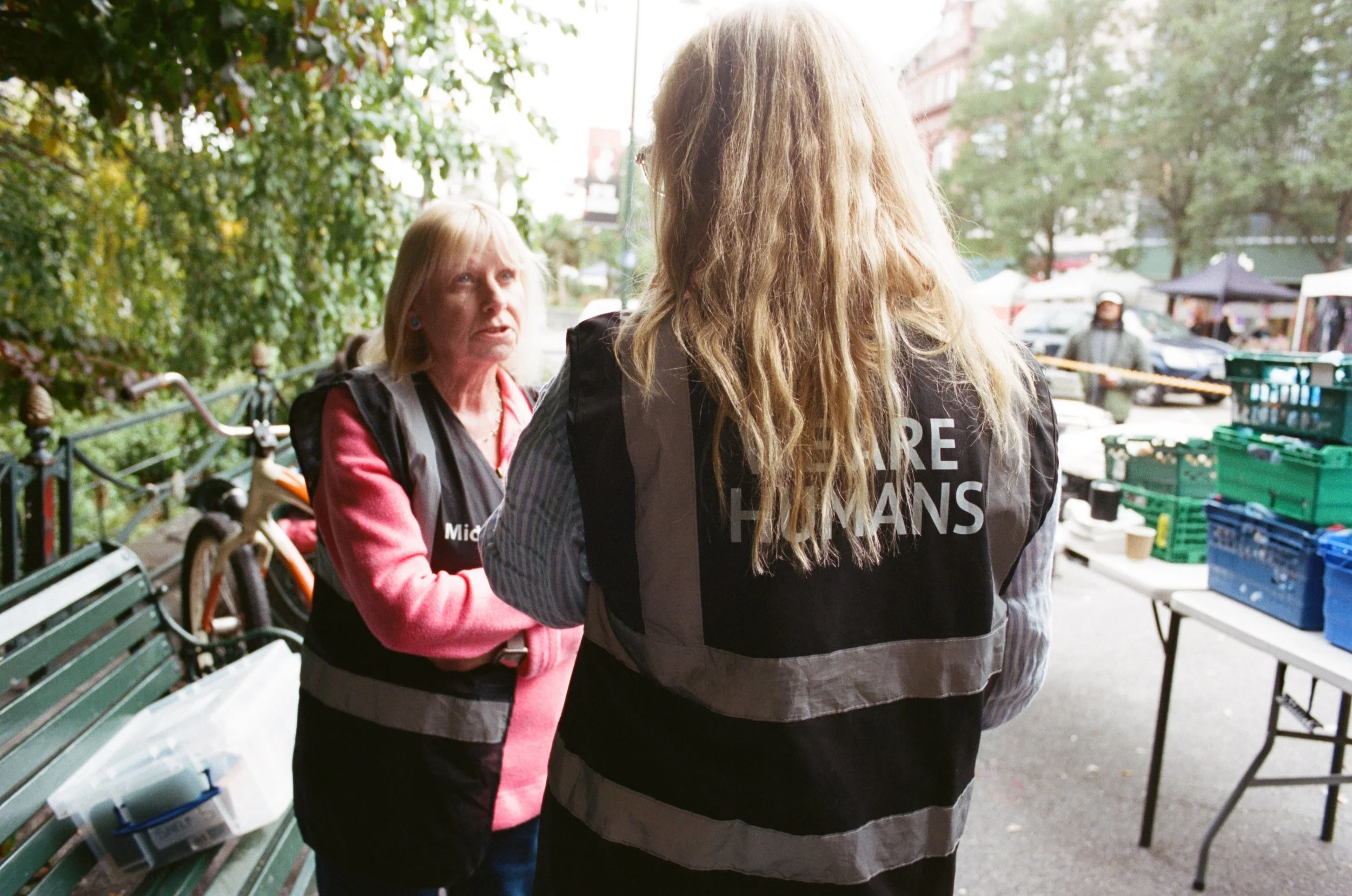 Two women engaging in conversation outdoors on a city street, with one woman facing away and the other woman facing forward, during daytime.