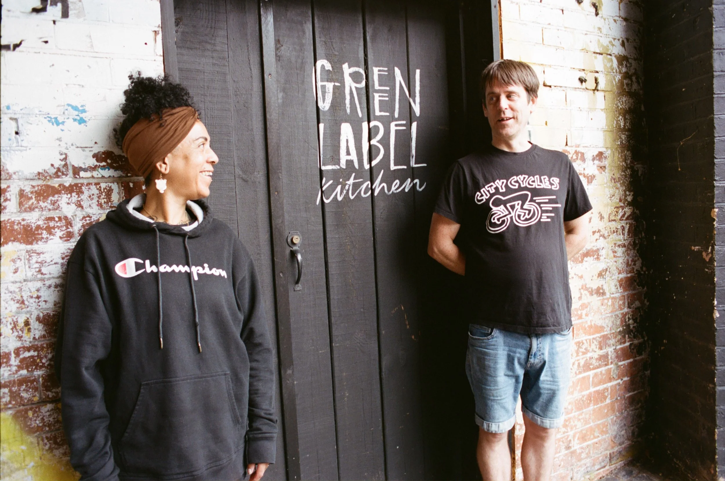 Portrait of two Green Label Kitchen team members standing beside the branded entrance door. Photo by Shahab Notghi.