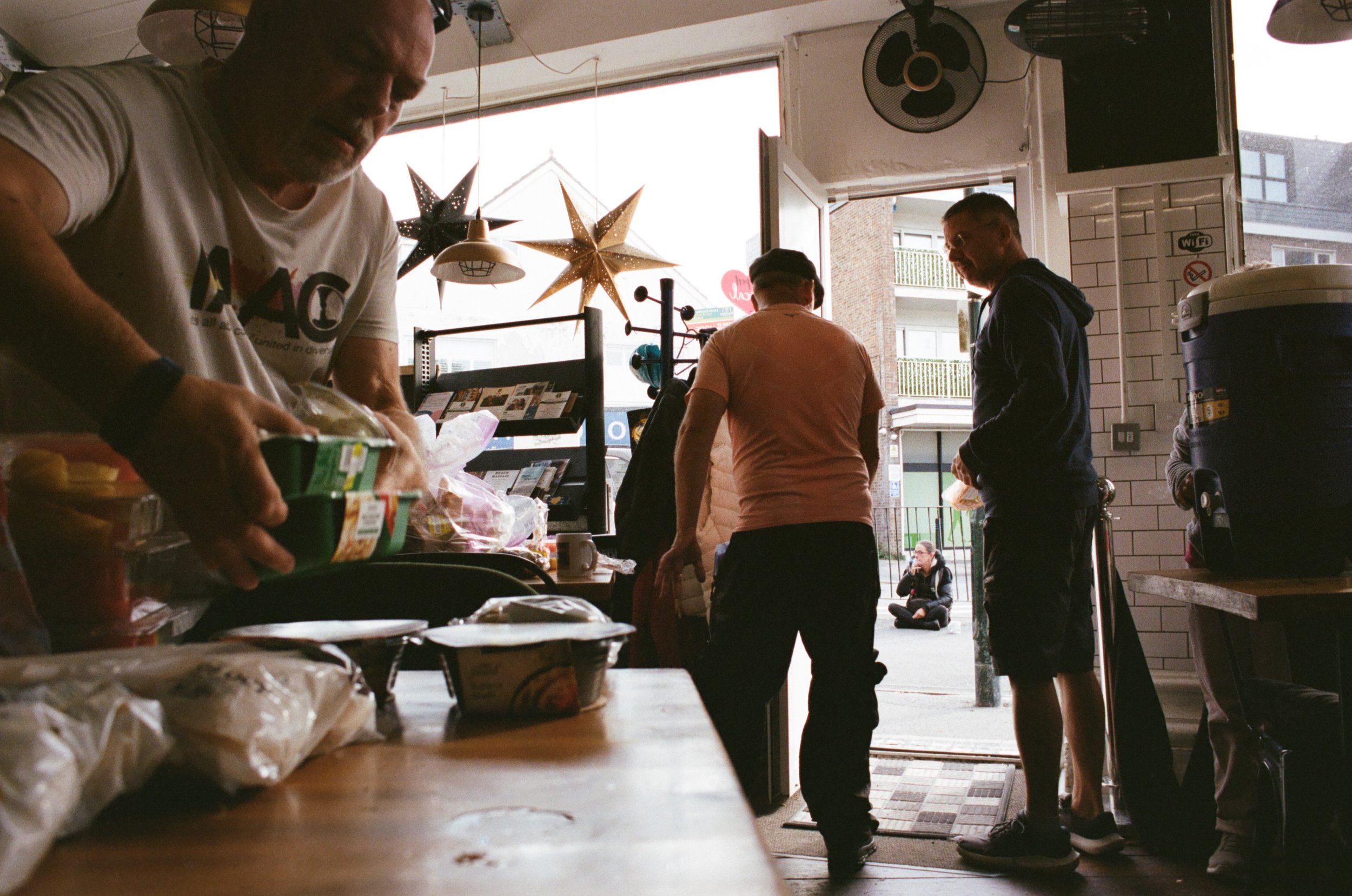 People inside a cafe or bakery, some ordering or paying at the counter, with star-shaped decorations hanging from the ceiling, and an open door leading outside where a woman is sitting on the sidewalk.