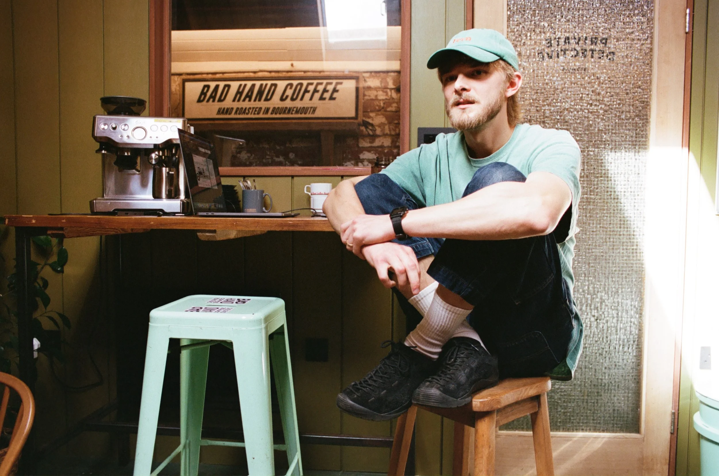 Portrait of a man in a green cap sitting beside an espresso machine and Bad Hand Coffee signage. Photo by Shahab Notghi.