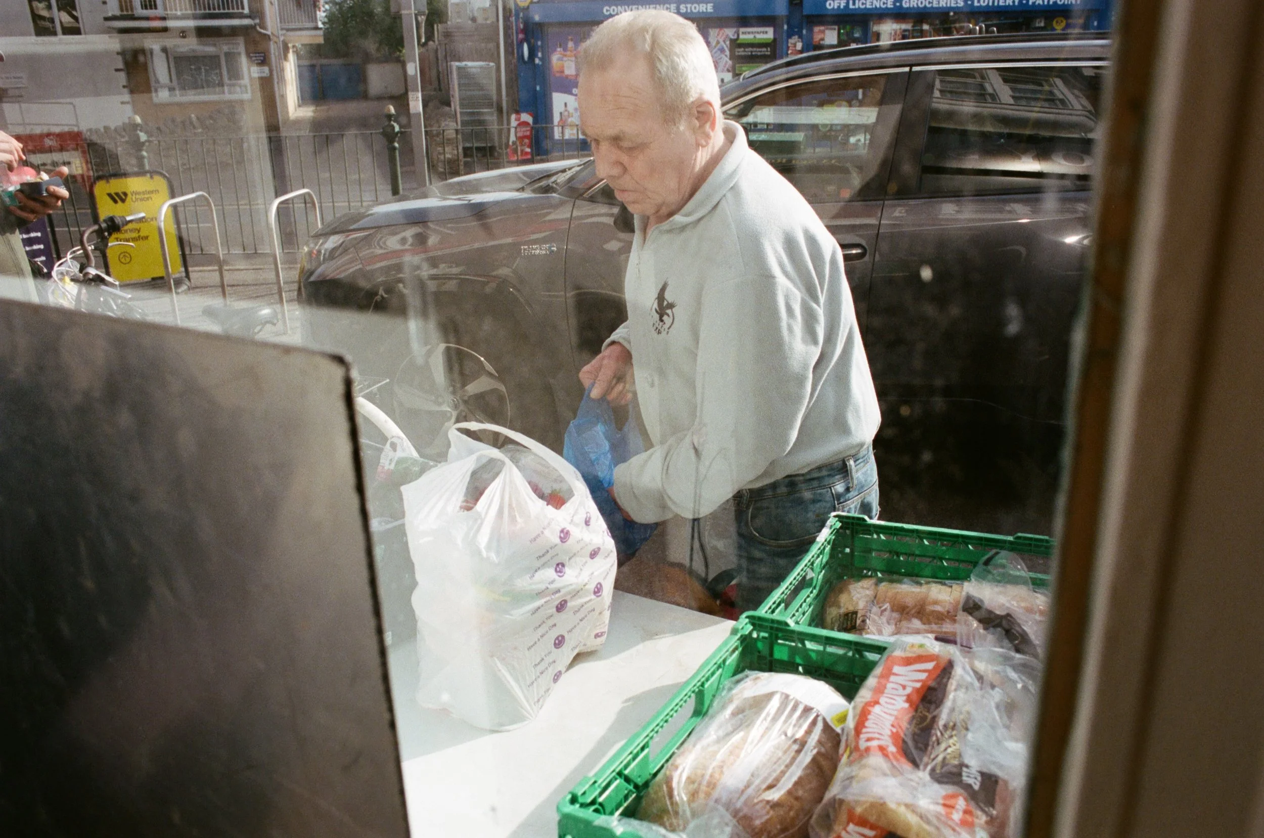 An elderly man with short white hair dressed in a light gray sweatshirt and jeans, standing inside a grocery store, appears to be shopping for bread and other items.