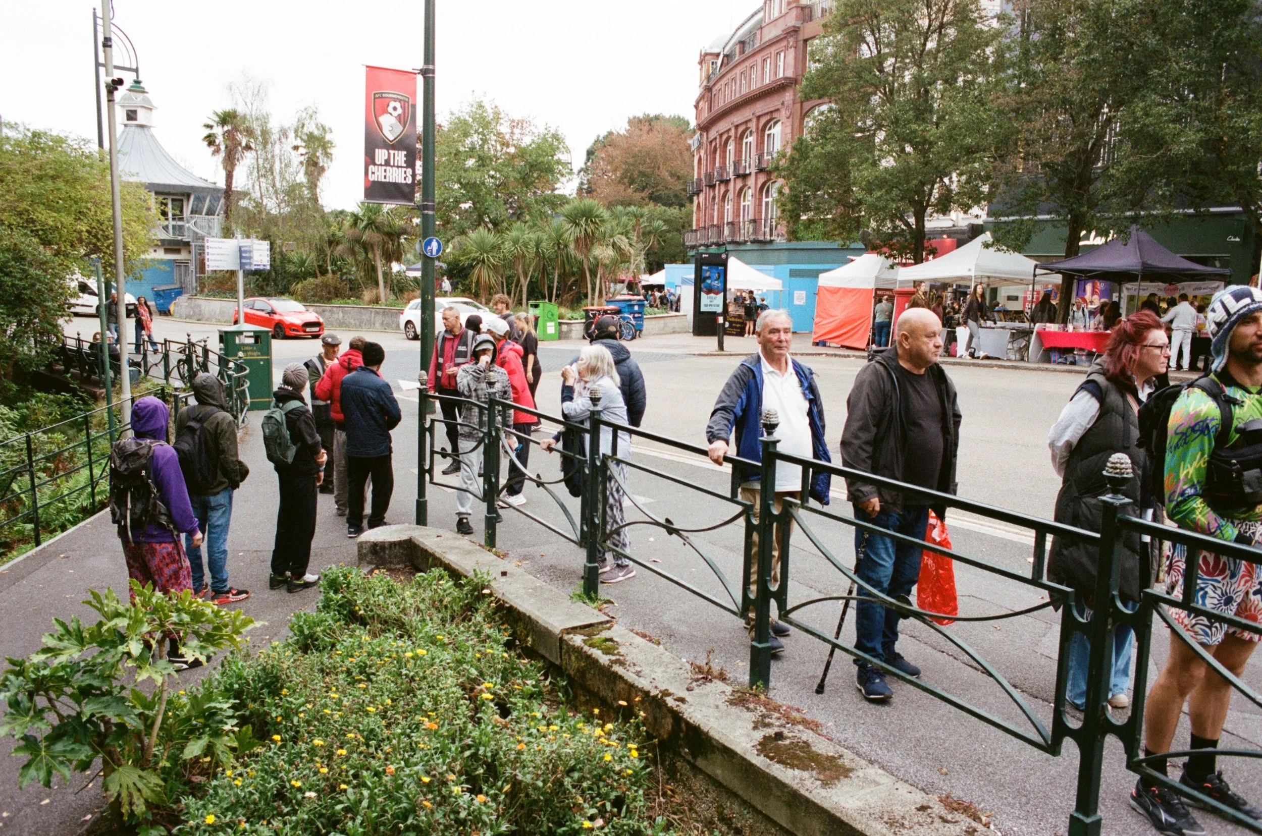 People walking and standing on a sidewalk with market stalls and tents in the background. There are trees and a pink brick building, with a traffic sign, cars, and a banner reading 'UP THE CHERRIES' in the scene.