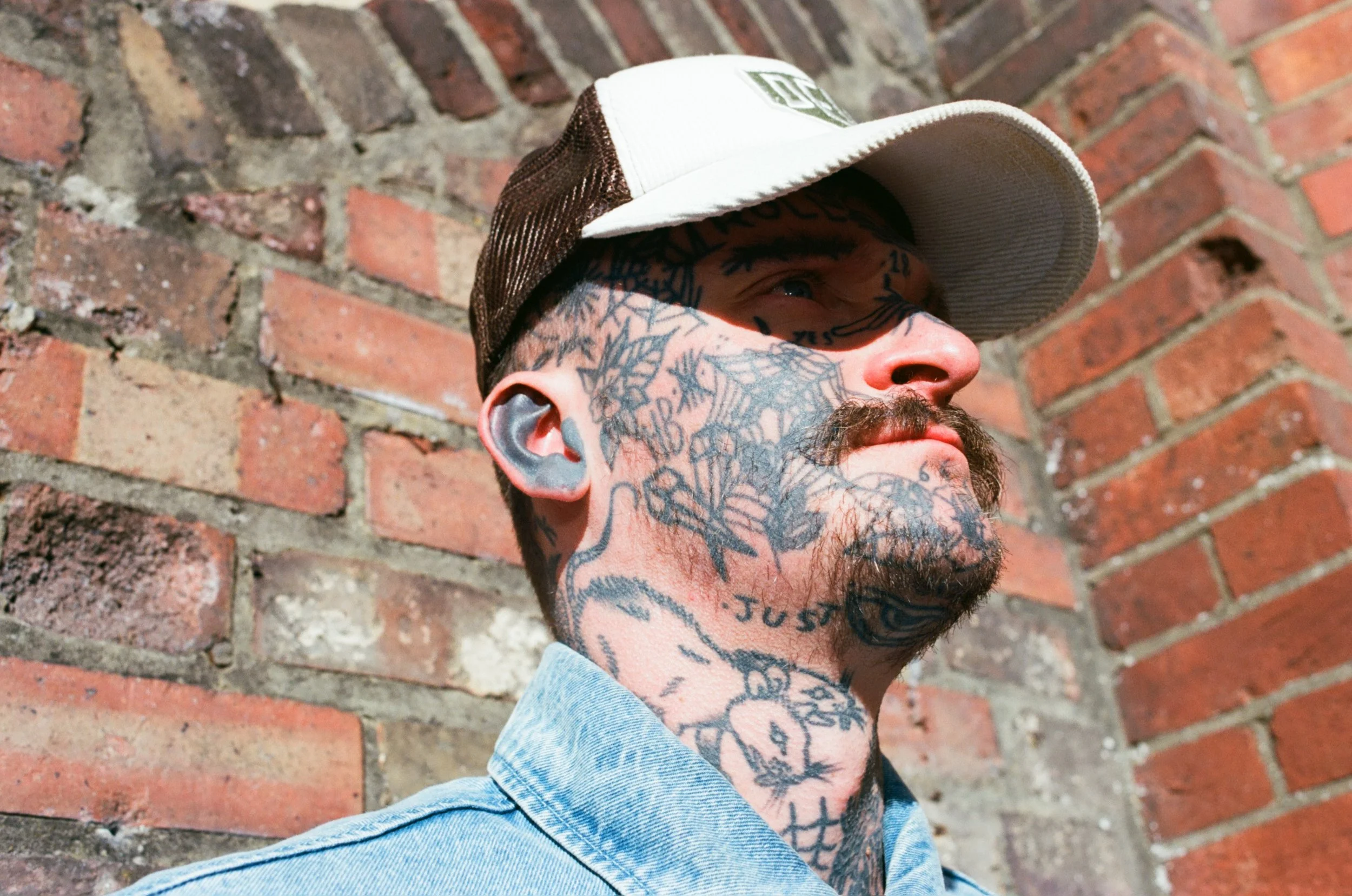 Close-up of a young man with tattoos on his face, neck, and head, wearing a white and brown baseball cap and denim jacket, standing against a red brick wall.