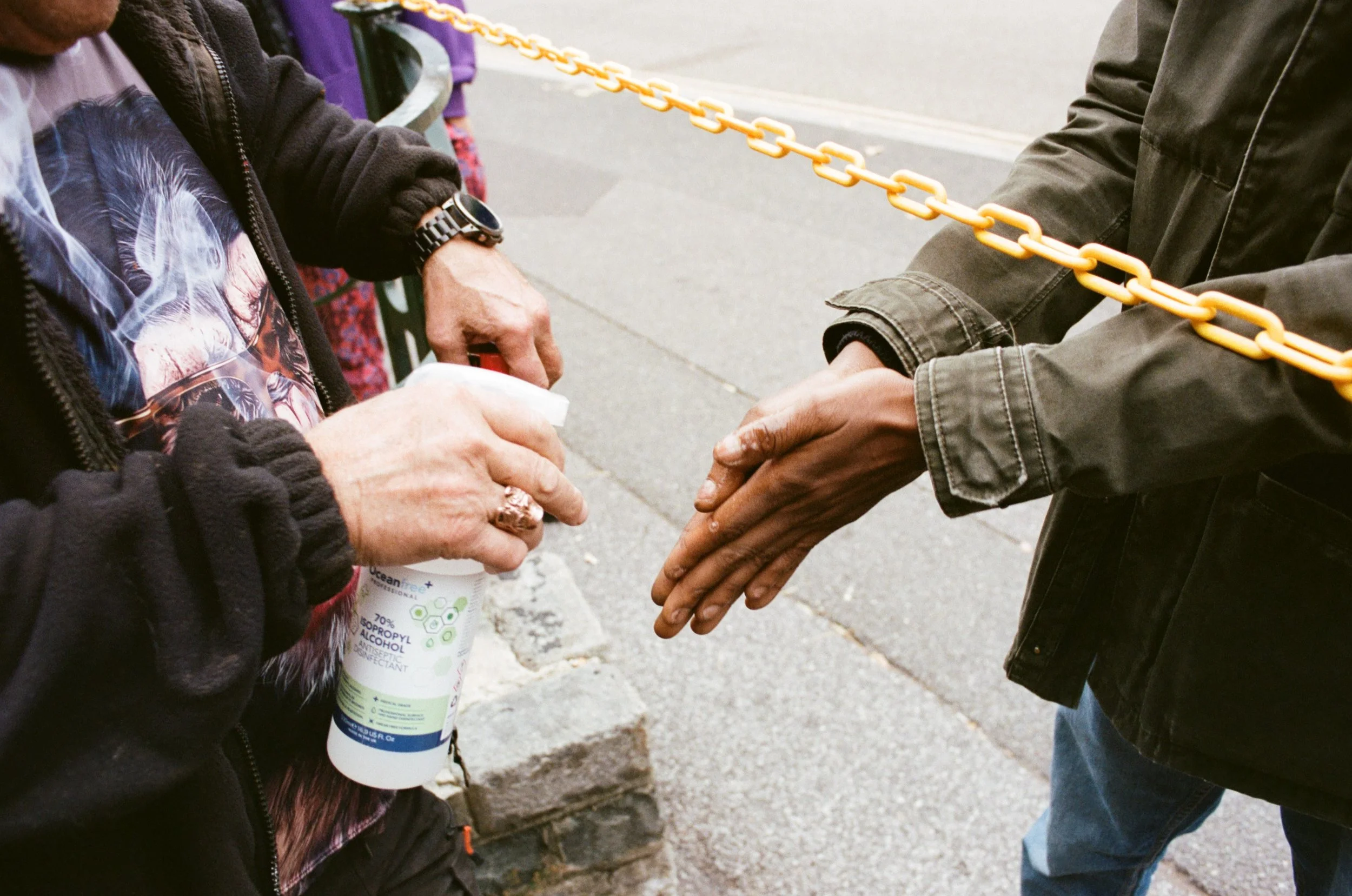 Two people are exchanging hand sanitizer outdoors, with one person holding a white bottle labeled 'Alcohol-Based Hand Sanitizer' and the other person's hand extended to receive it. The scene includes a chain barrier and a sidewalk.
