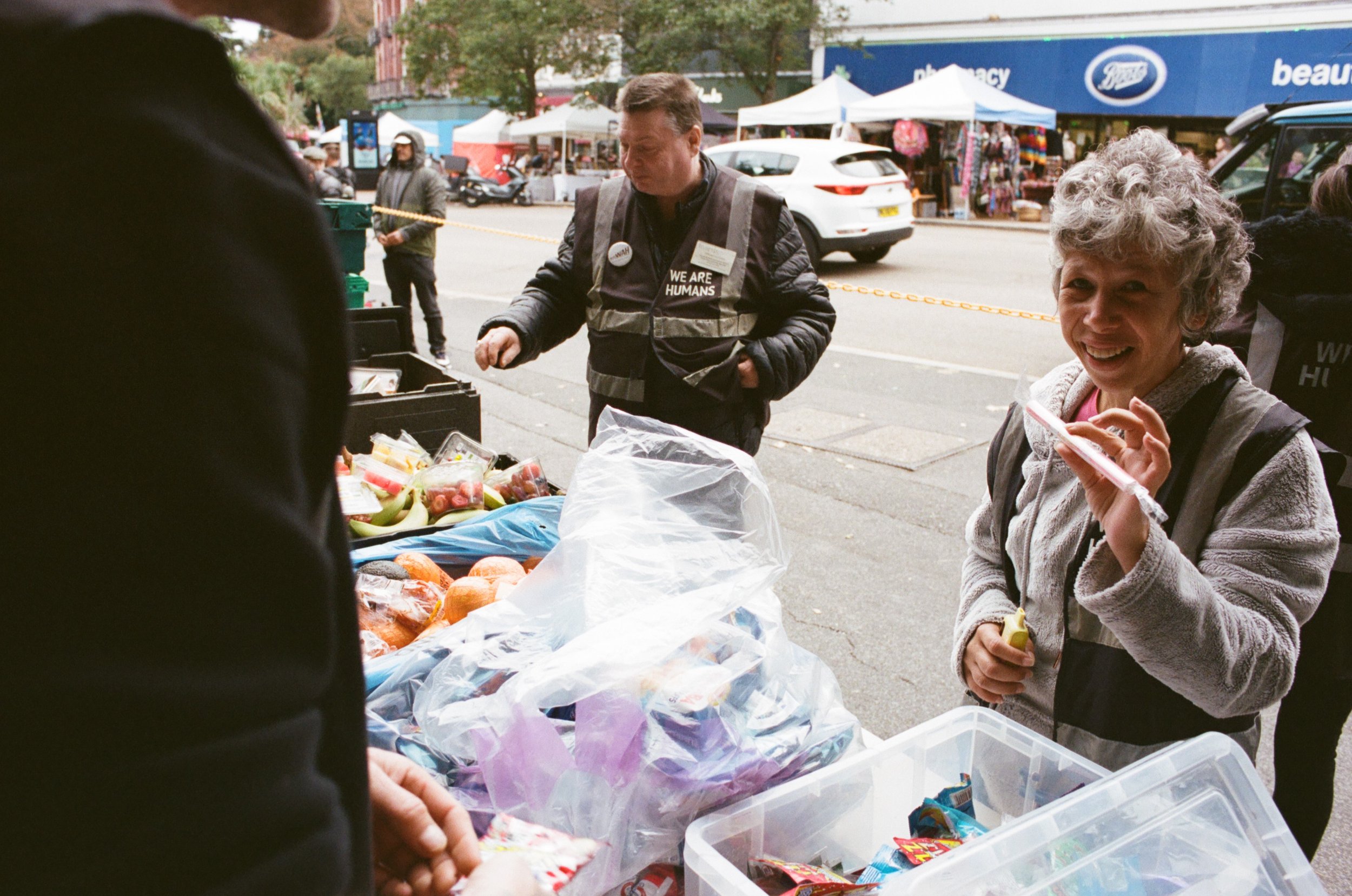 A woman and a man at an outdoor market stand selling candy and produce, with tents and buildings in the background.