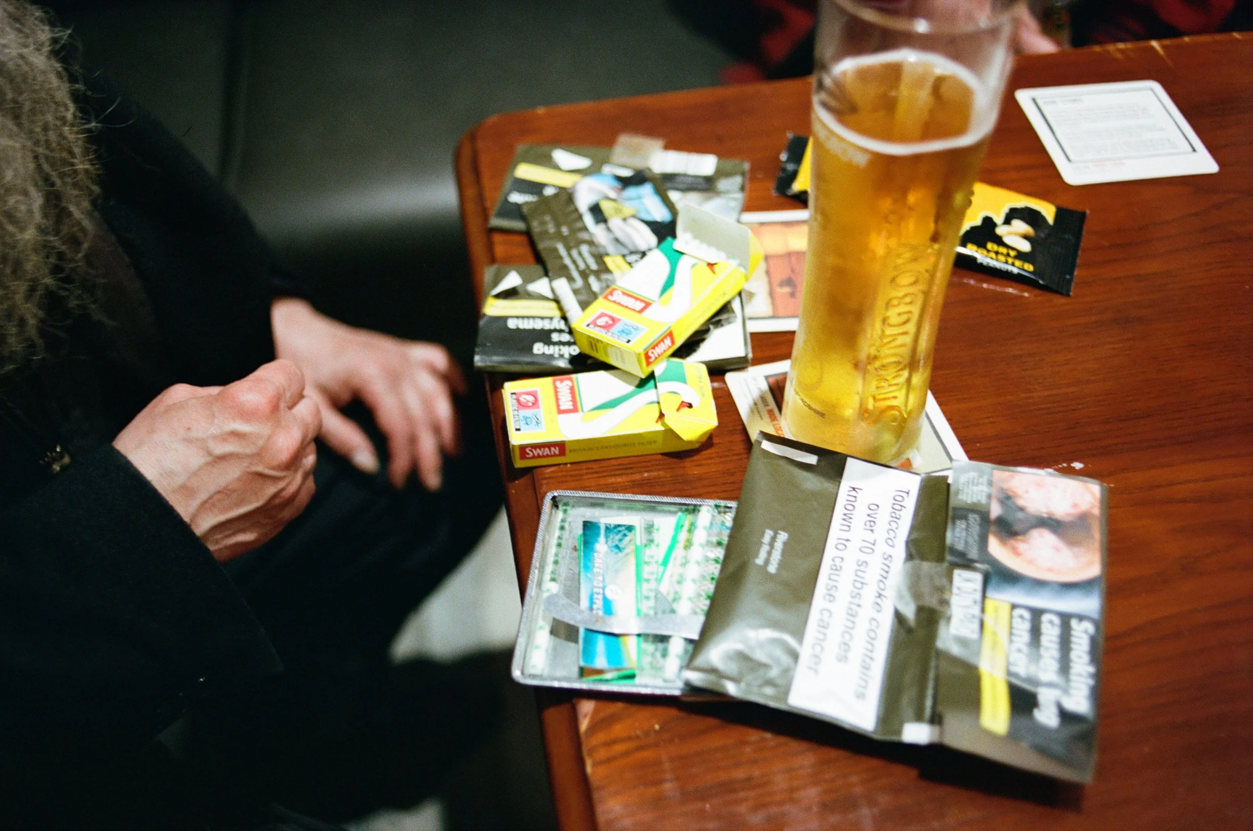 A person sitting at a wooden table, with a glass of beer in front of them and various packets of chewing tobacco and cards scattered on the table.