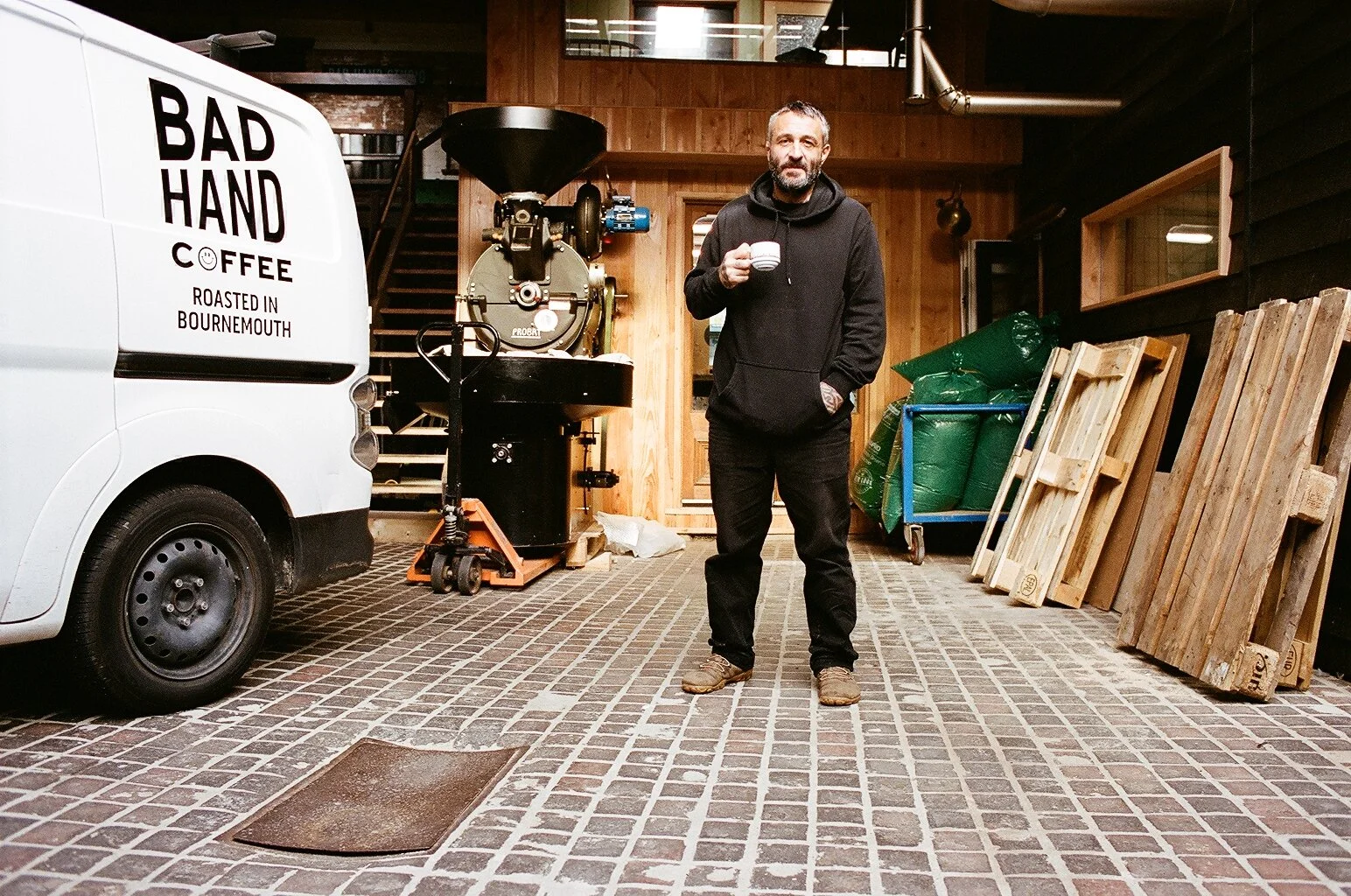 Bad Hand Coffee roastery in Bournemouth, with a coffee roaster and branded delivery van beside a man holding a cup of coffee. Photo by Shahab Notghi.