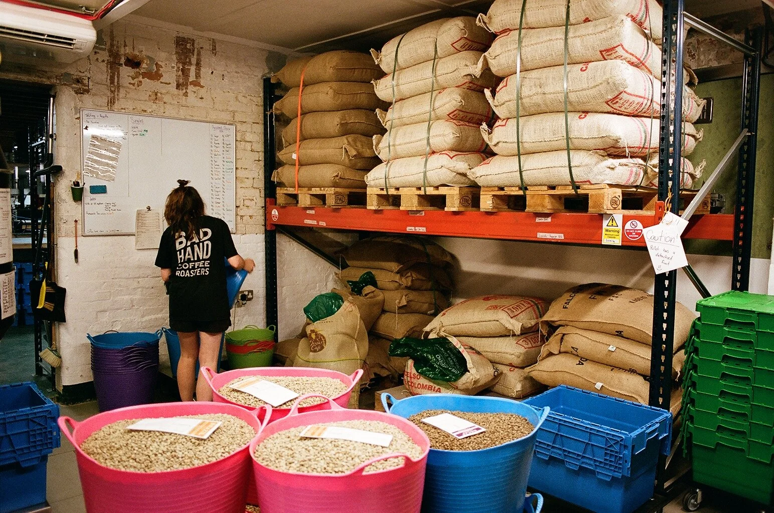 Green coffee beans displayed in colourful tubs beneath shelves of stacked coffee sacks at Bad Hand Coffee. Photo by Shahab Notghi.