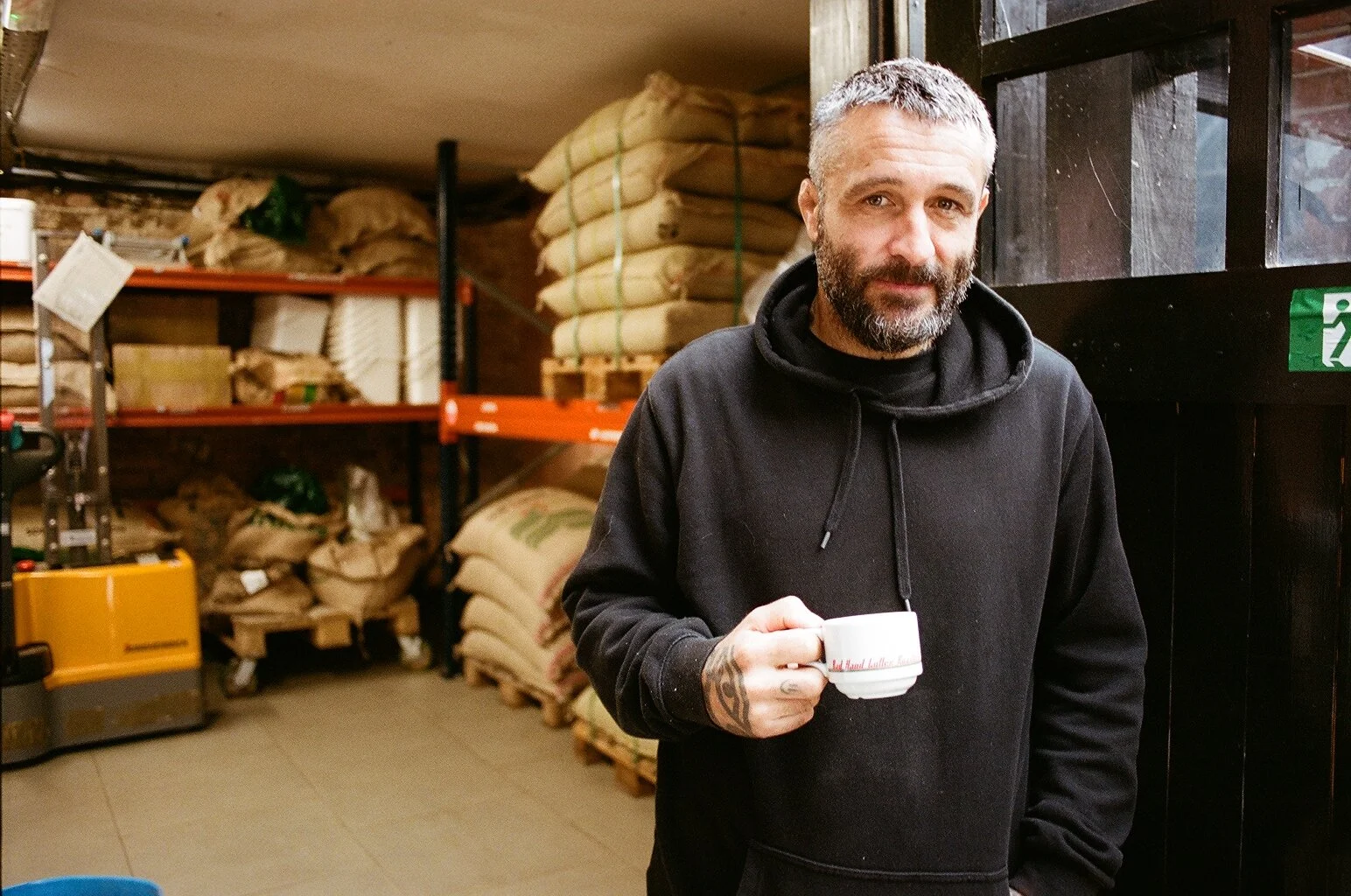 Portrait of a Bad Hand Coffee team member holding a cup of coffee in front of stacked coffee bean sacks. Photo by Shahab Notghi.