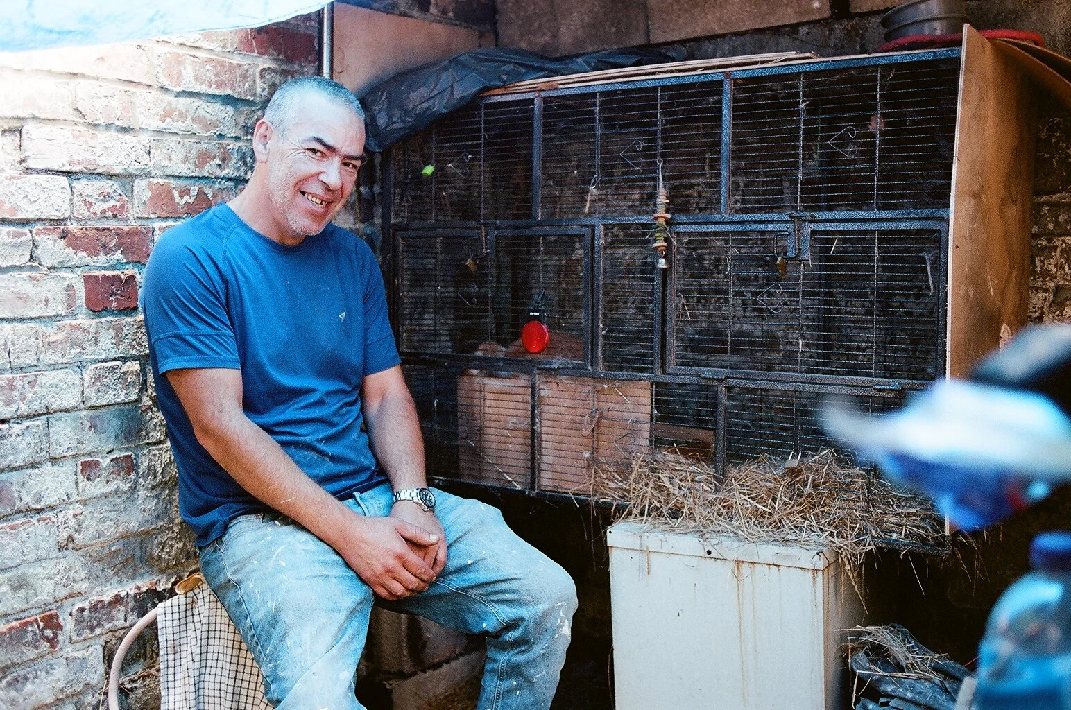A man with a shaved head and wearing a blue t-shirt, sitting next to a large birdcage with straw on the floor inside, in an indoor setting with brick walls.
