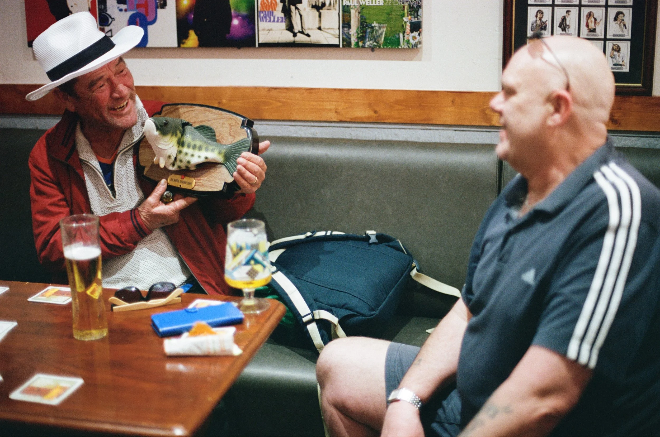 A man wearing a white fedora hat and red jacket holding a fish-shaped trophy, smiling at another man in a gray Adidas shirt, in a setting with a wooden wall, colorful artwork, and framed photographs on the walls. There are drinks and game items on th