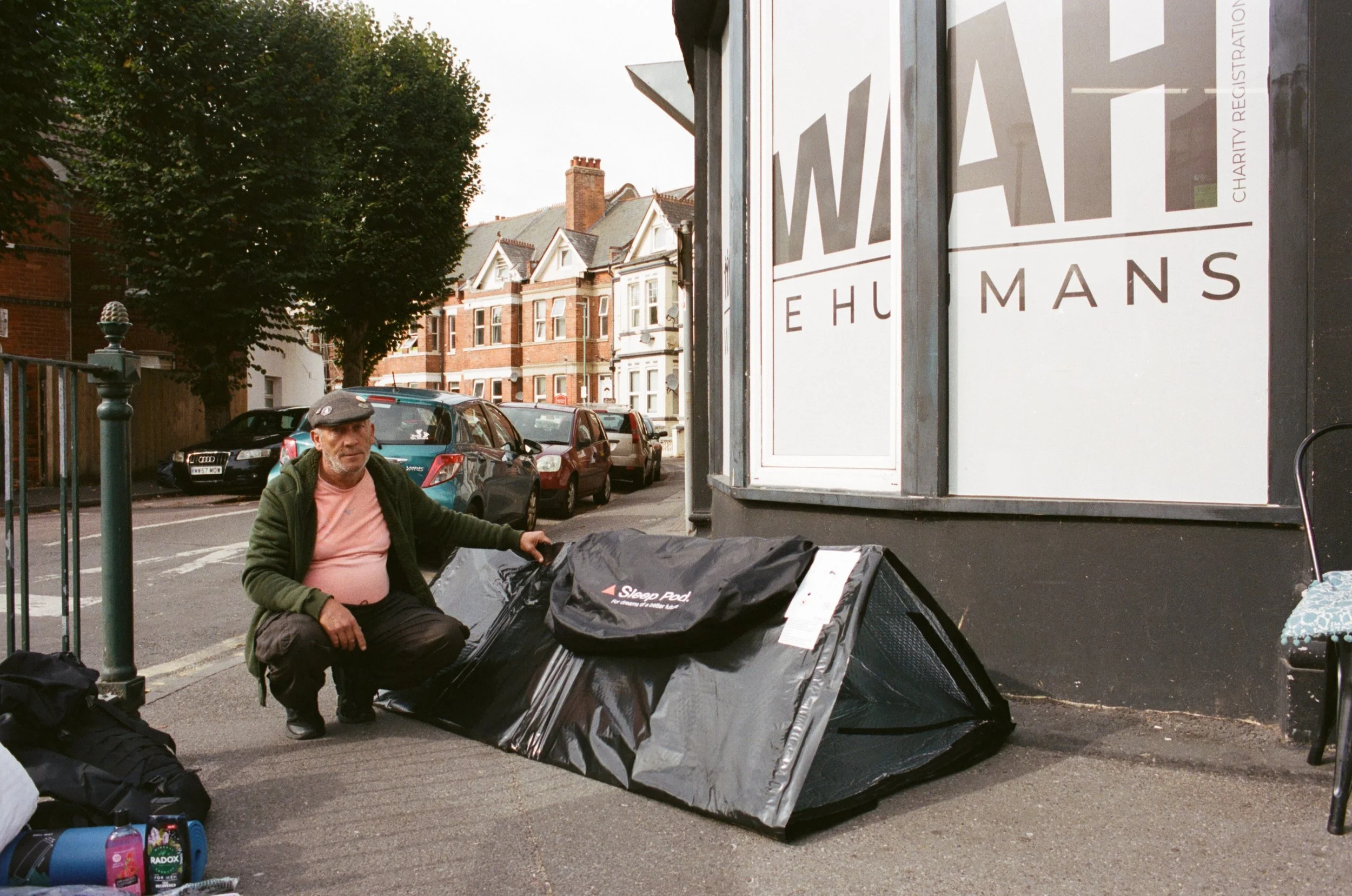 A man is sitting on the sidewalk in front of a storefront, surrounded by black trash bags. There are parked cars and brick houses in the background, and a tree with green leaves. A sign on the storefront displays the words 'WAI' and 'EHUMANS'.