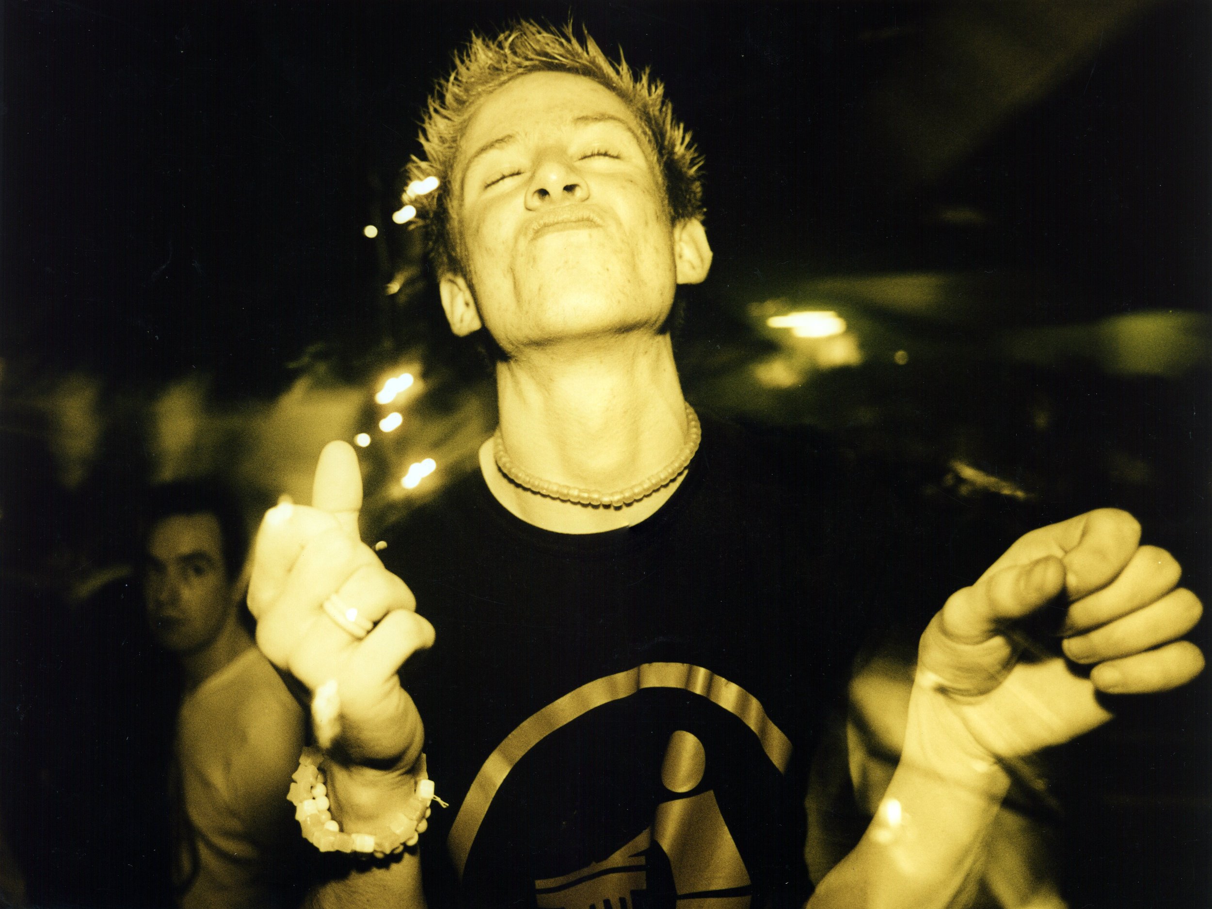 A person with short spiky hair, smiling with eyes closed, wearing multiple necklaces and bracelets, and holding their hands up with fingers curled, in a dark environment with blurred lights in the background.