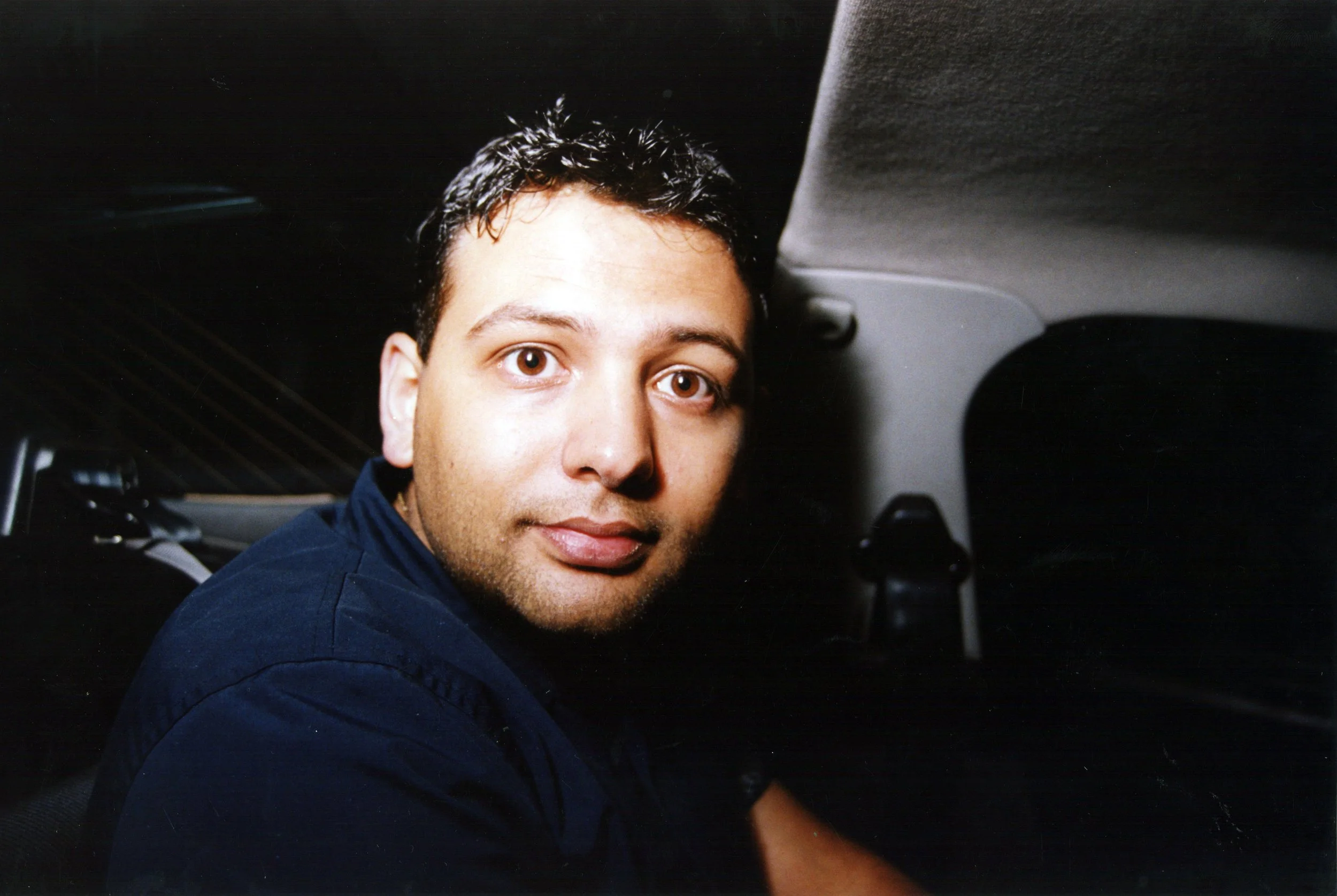 A young man with short, dark, wet-looking hair and brown eyes sitting inside a vehicle, wearing a dark blue jacket.