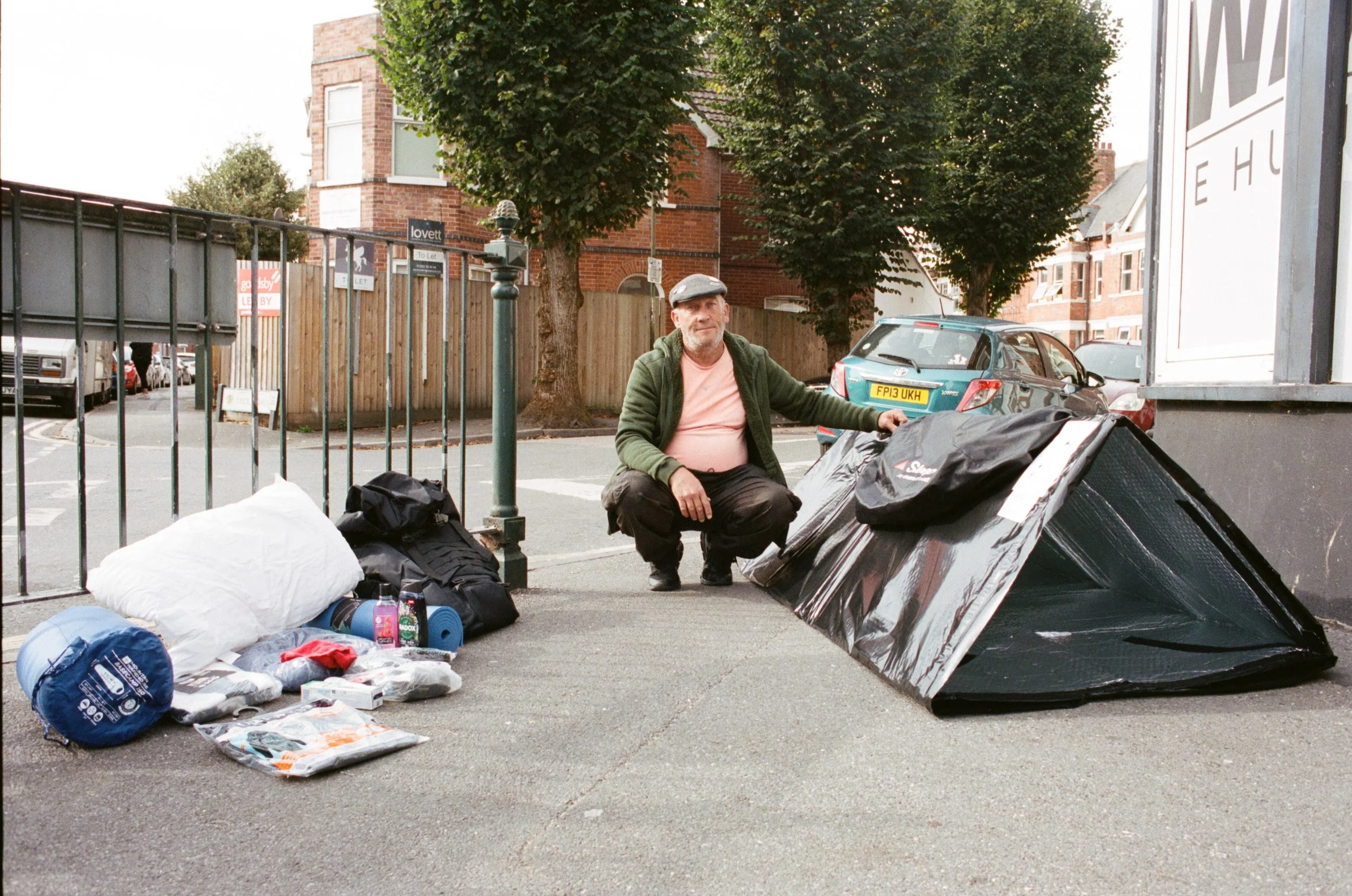 A man with a beard and gray cap squatting on the street near trash bags, boxes, and belongings, with trees, brick buildings, and parked cars in the background.