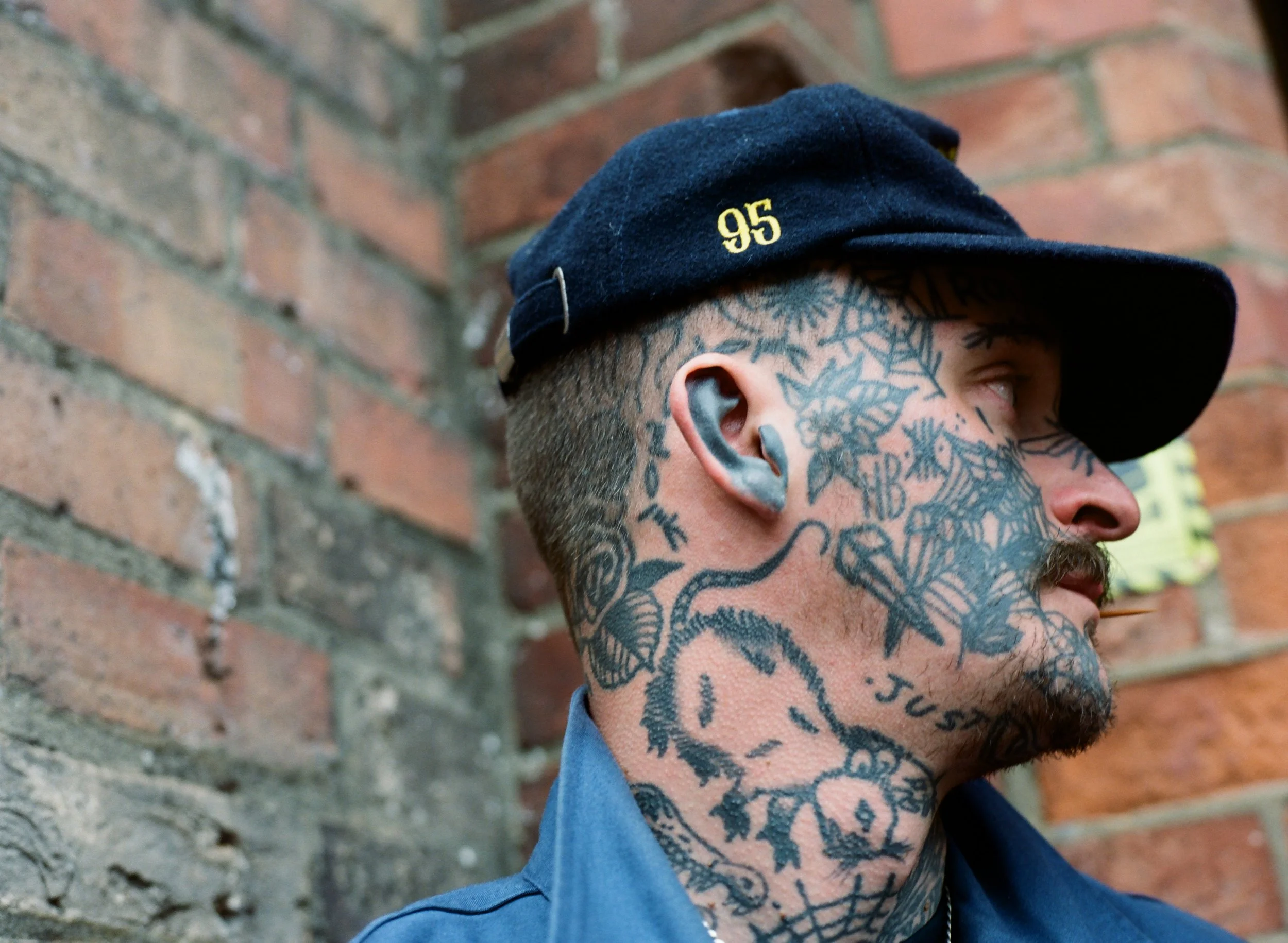 Close-up of a man with tattoos on his neck and face, wearing a navy blue cap with the number 95 embroidered in yellow, standing against a brick wall.