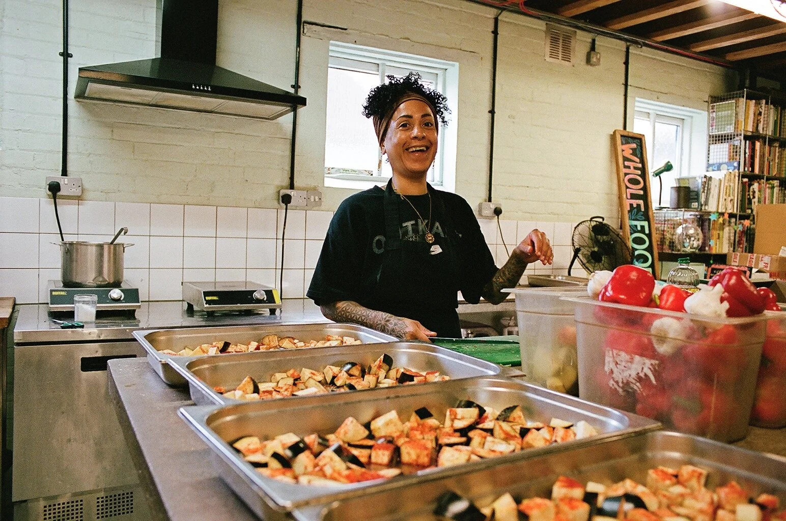 A woman with tattoos wearing a black shirt and apron stands in a kitchen, smiling at the camera. There are trays of chopped vegetables, likely eggplant and tomatoes, and containers of peppers or red and white peppers on the counter. The background sh