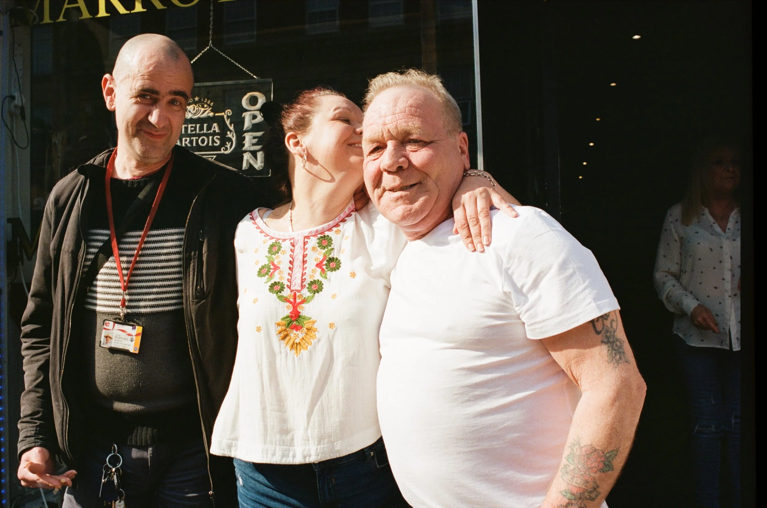 Three people smiling and posing together outside a building. One woman is embracing a man, and another man is standing to the side. The woman is wearing a traditional embroidered blouse, and the man she is hugging has tattoos on his arm.