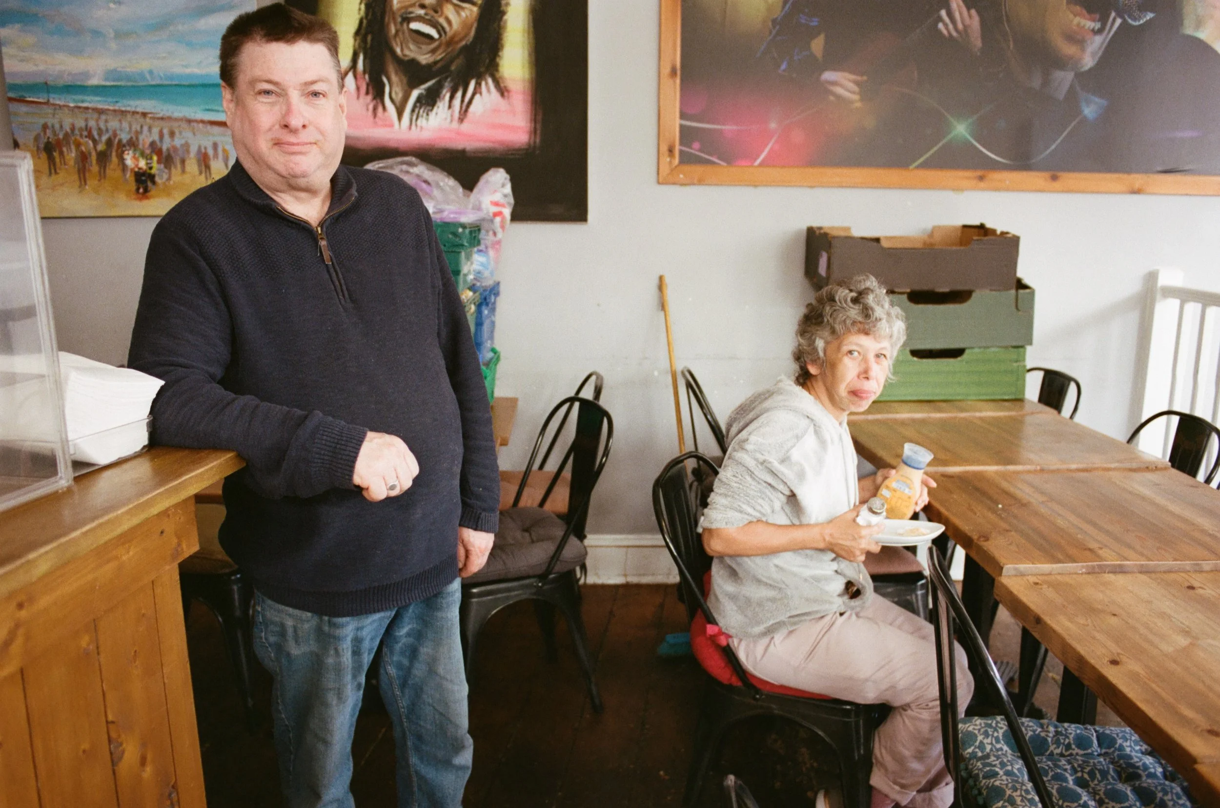 A man standing next to a seated woman at a wooden table inside a room with colorful artwork on the wall. The man is wearing a black zip-up sweater and jeans, and the woman is eating breakfast with a bottle of orange juice in her hand. The woman has c
