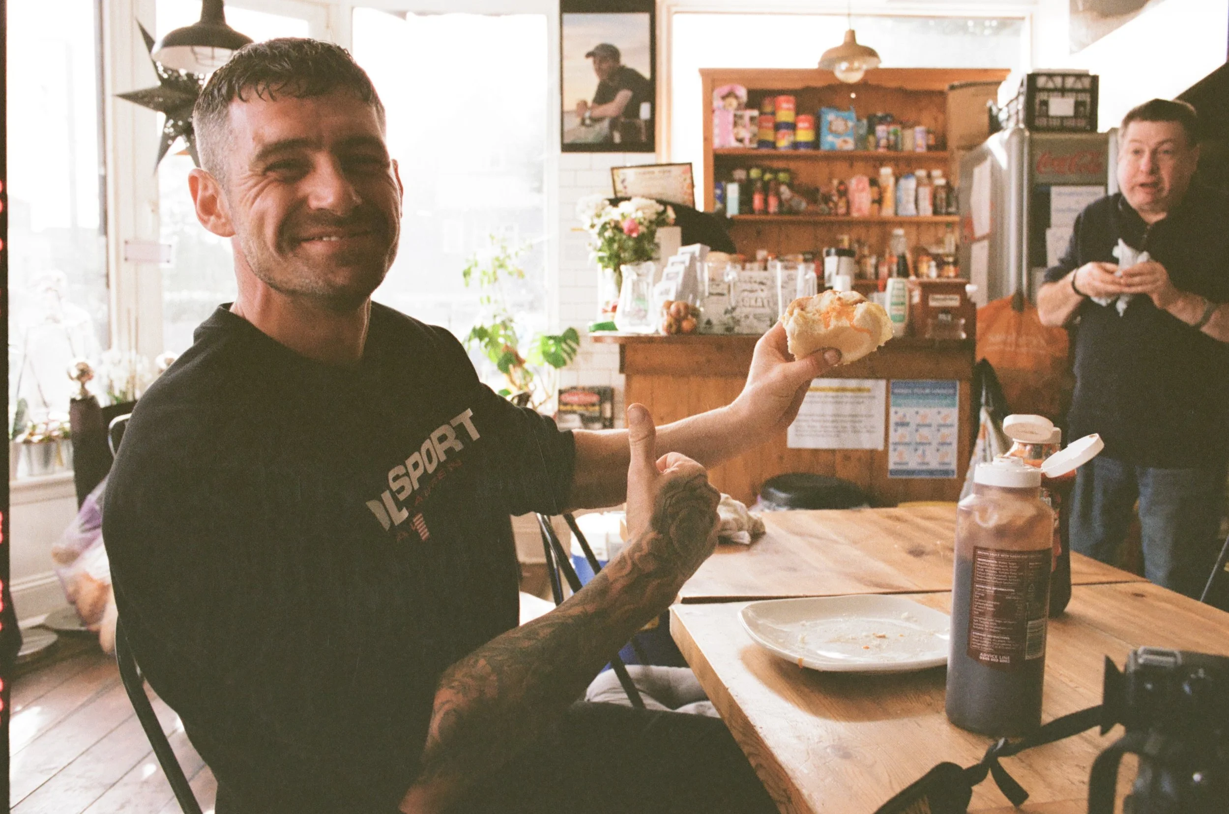 A man with tattoos and a black T-shirt sitting at a wooden table in a cozy cafe, smiling and holding a piece of bread or pastry. Behind him, a woman with short dark hair is holding a napkin or tissue near a counter filled with various food items and 