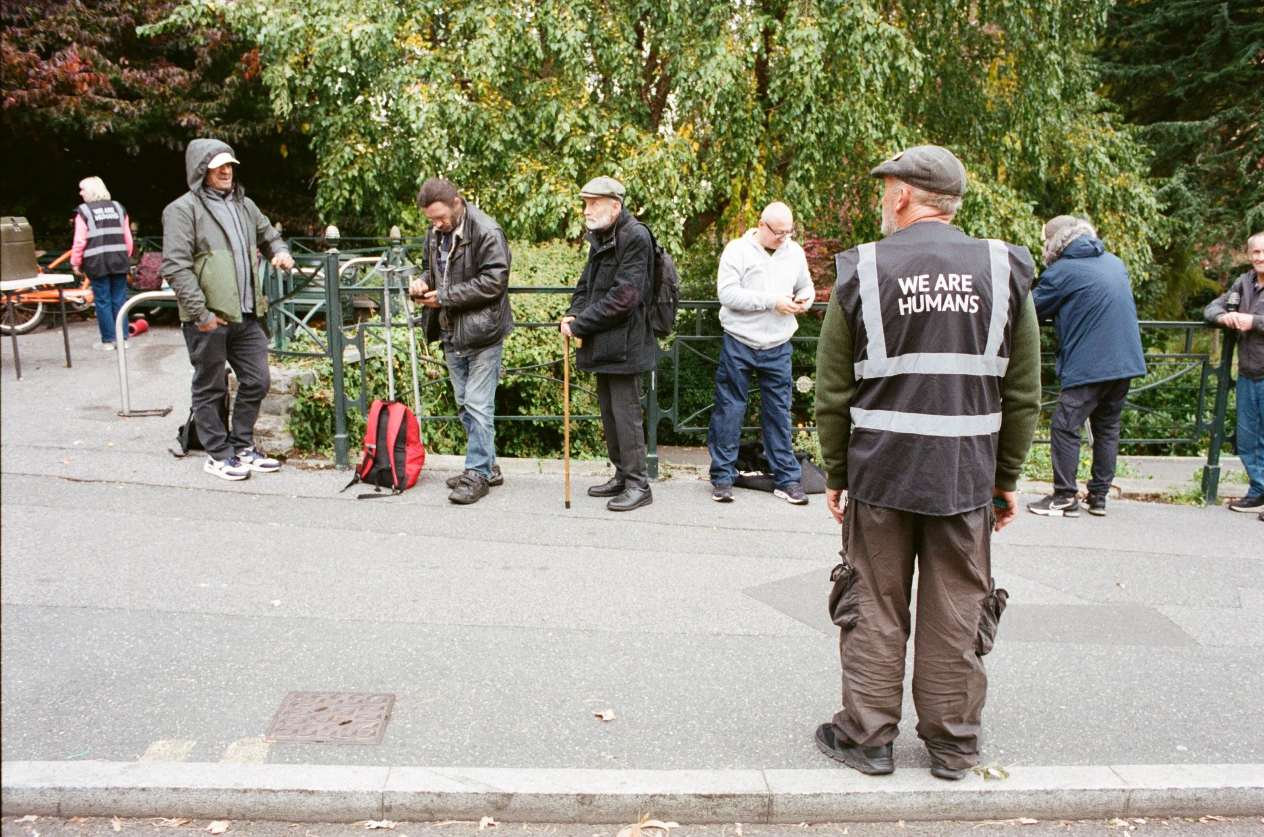 A group of people standing outdoors on a sidewalk near a green railing and trees, some looking at their phones, one person wearing a vest with the words 'WE ARE HUMANS' on the back.