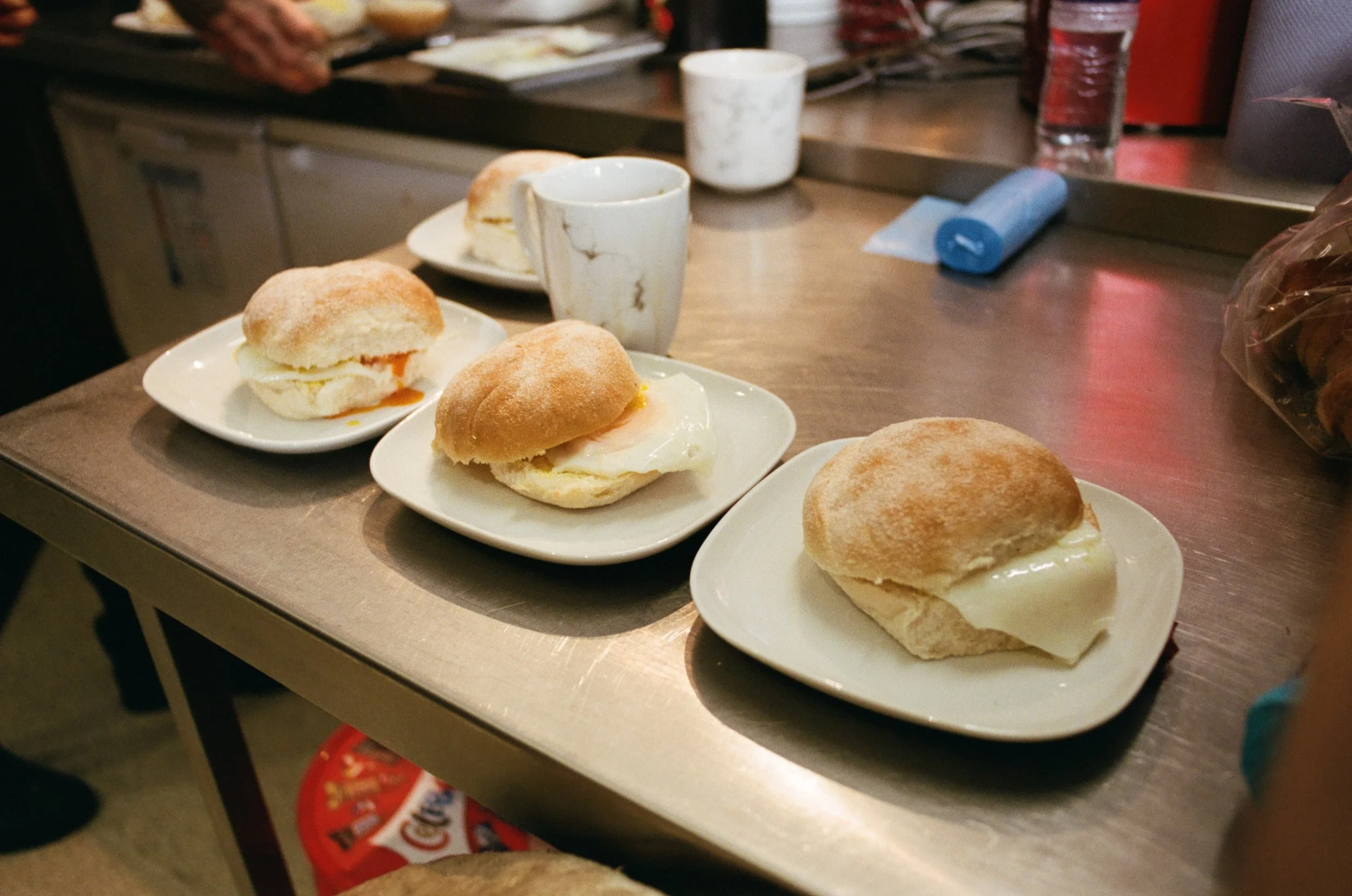 Four plates with breakfast sandwiches featuring fried eggs and English muffins on a counter at a cafe or diner. Two cups and a glass of water are also visible.
