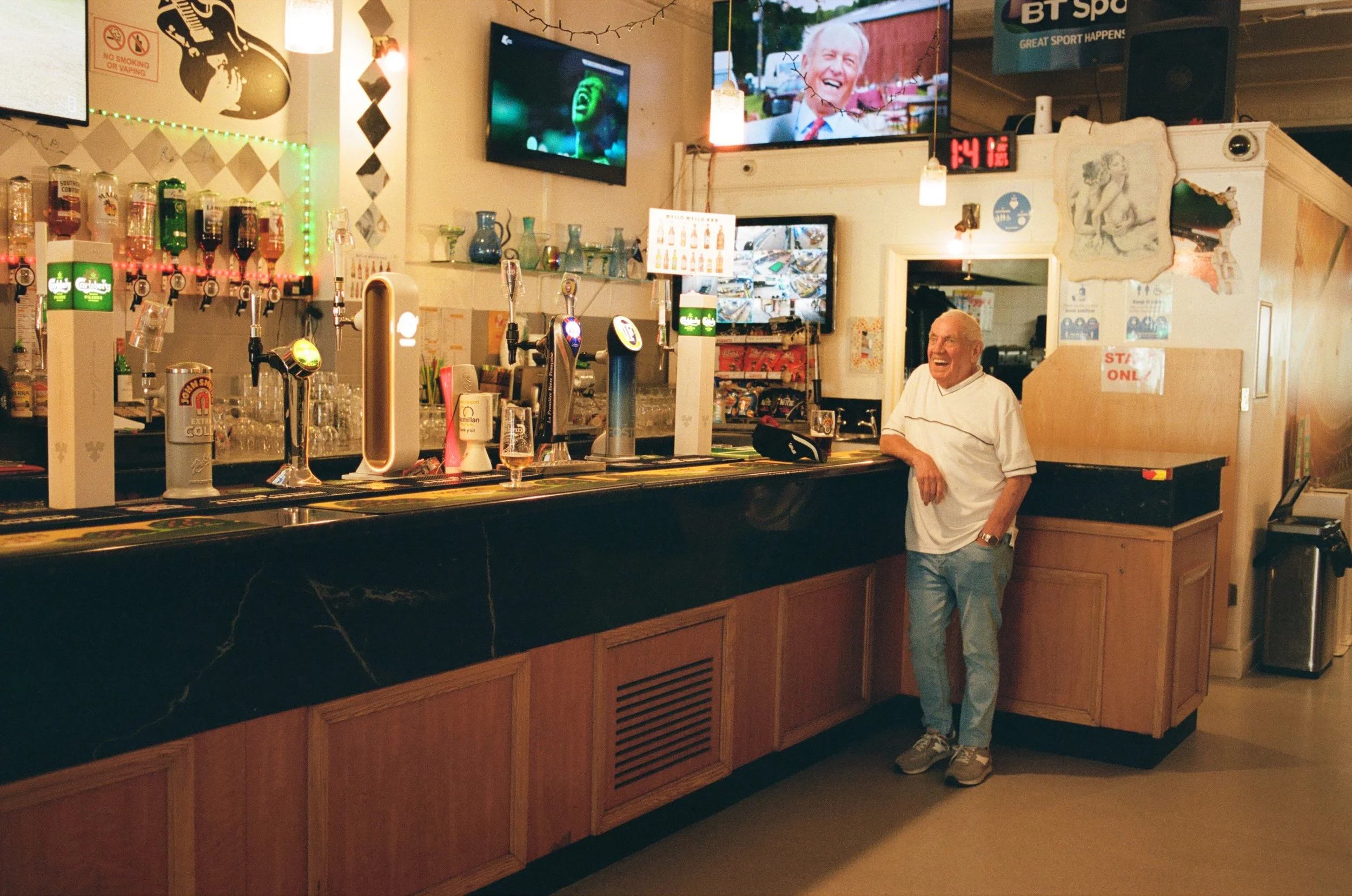 An older man standing at a bar counter, smiling. The bar has beer taps, glassware, and various bottles. There are multiple television screens mounted on the wall, showing different images. The decor includes string lights and art on the walls. The ma