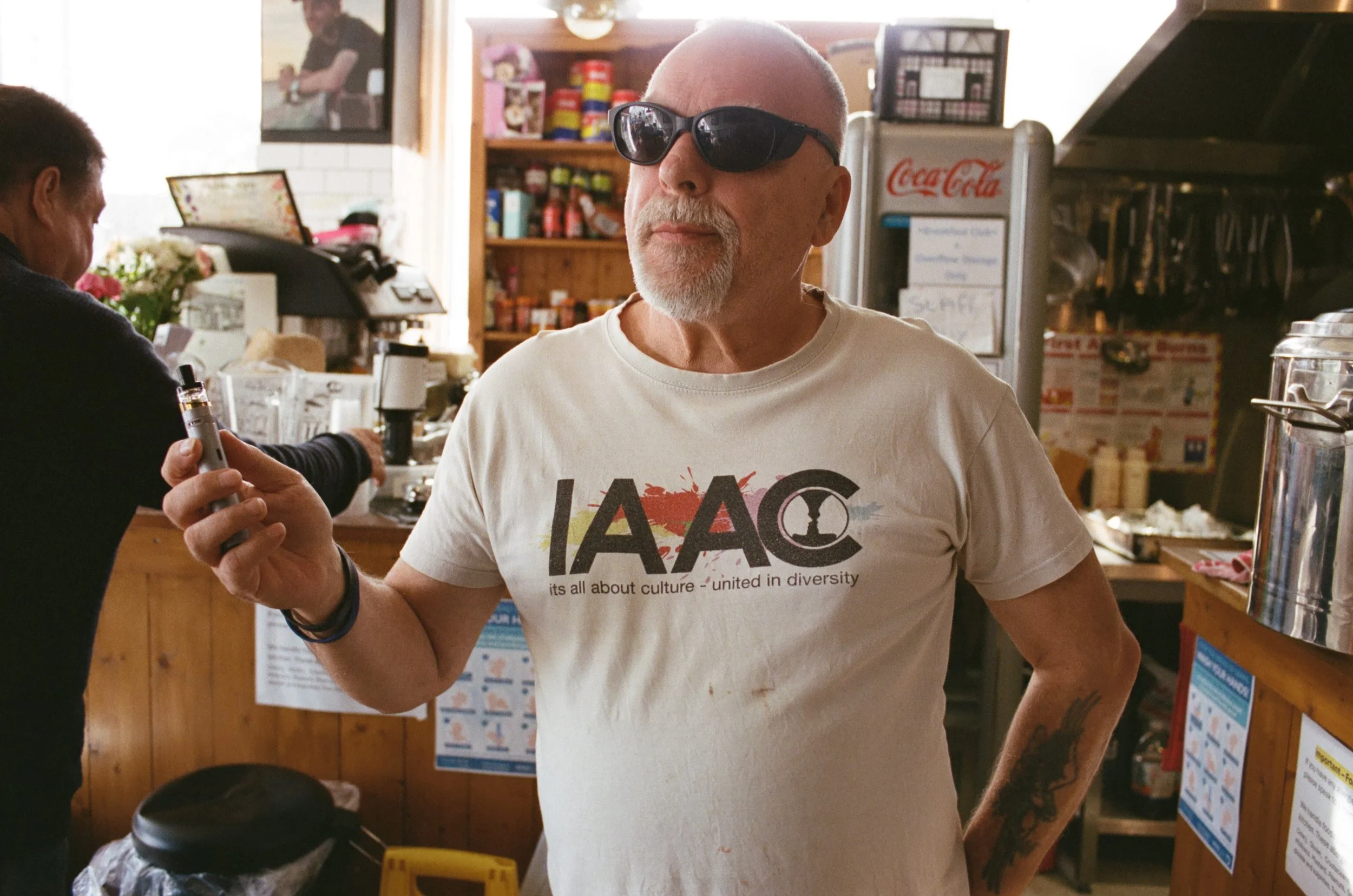 A man with a white beard, wearing sunglasses and a white T-shirt that reads 'LACC' and 'it's all about culture - united in diversity,' is standing inside a cafe or small shop. He is holding a vape pen in his right hand and has a tattoo on his left fo