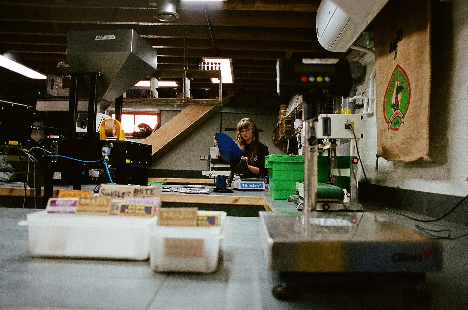 Coffee production workspace with industrial machinery, weighing equipment and a team member handling green coffee beans. Photo by Shahab Notghi.