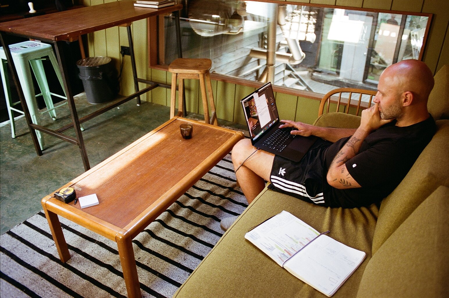 Man working on a laptop in the Bad Hand Coffee tasting room, surrounded by wooden tables, stools and large windows. Photo by Shahab Notghi.