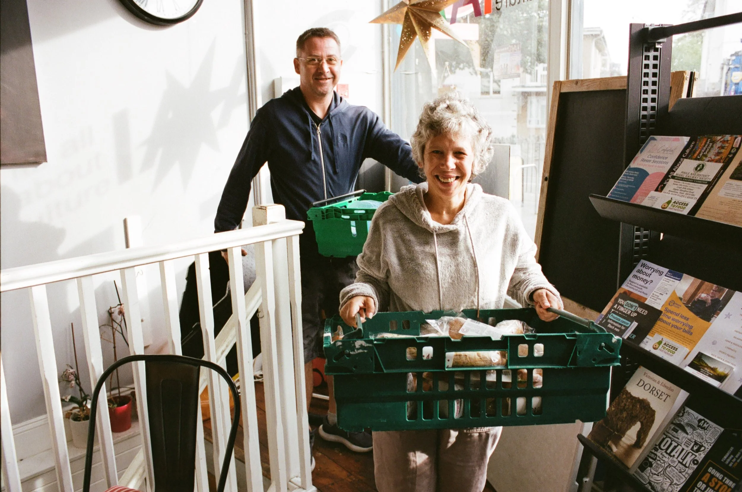 A woman standing behind a shopping cart with a man standing next to her in a bookstore or cafe. The woman is smiling and looking at the camera, wearing a gray hoodie, and the man is smiling, wearing glasses and a dark jacket. They are near books and 