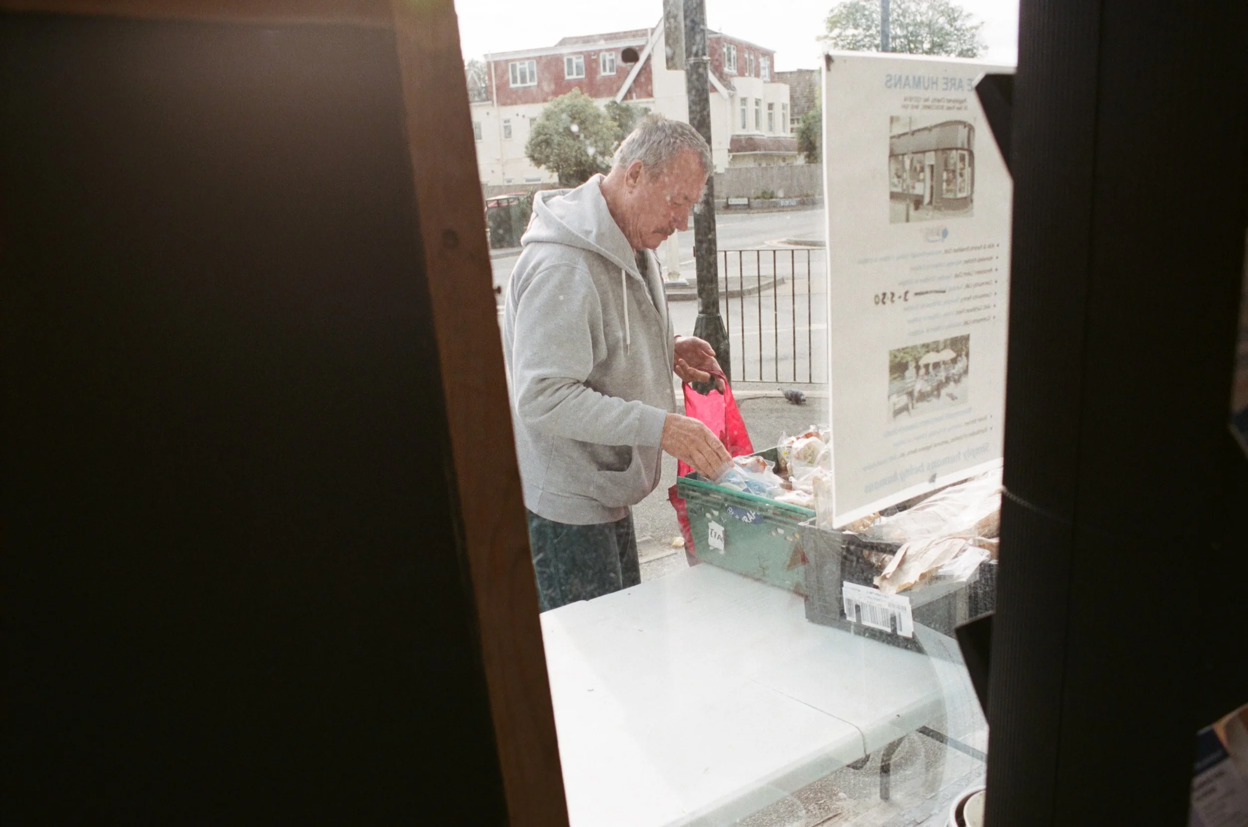 Older man in gray hoodie looking at a bag of groceries outside a store, viewed through a gap or opening in the store window.
