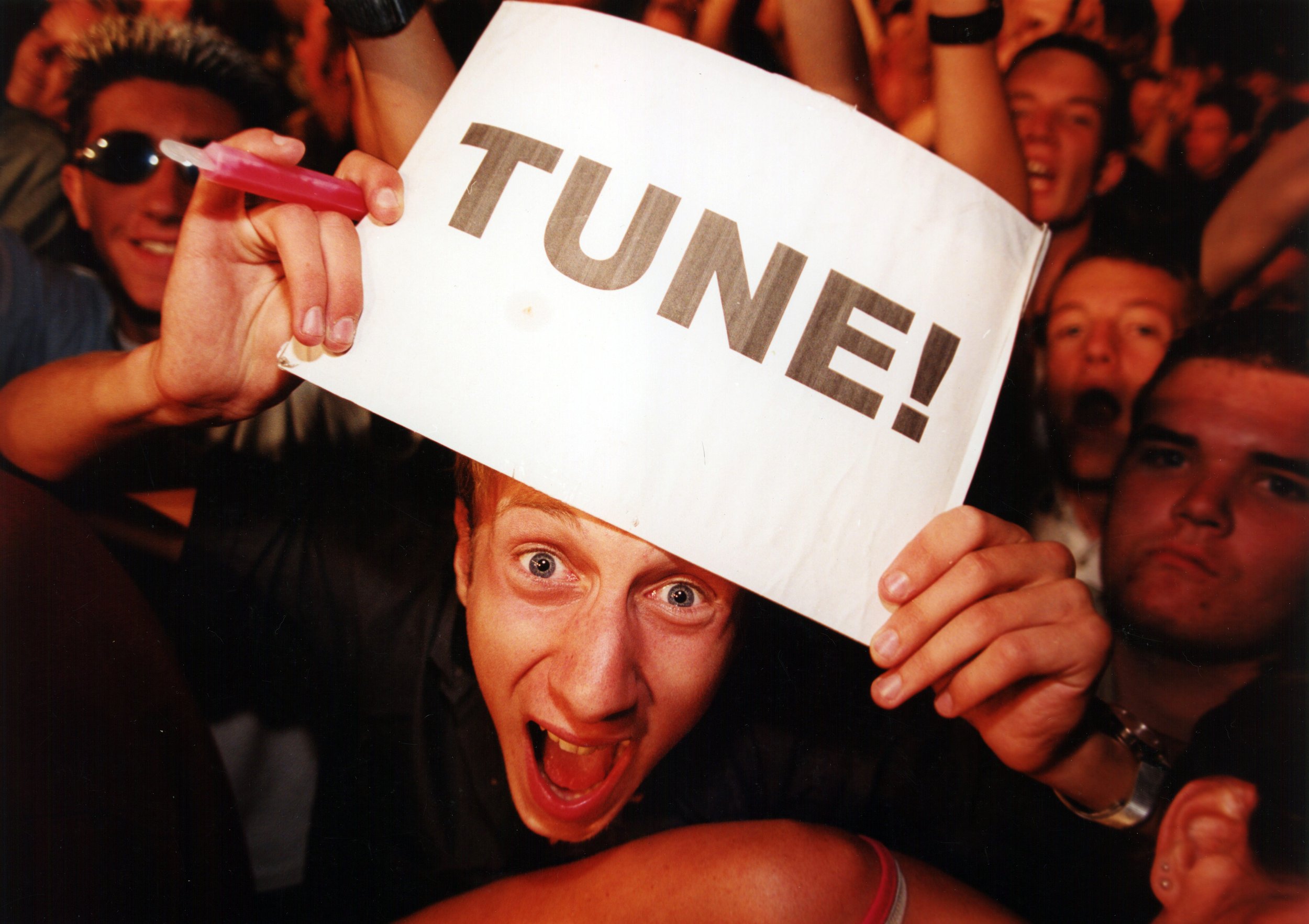 Excited woman with open mouth, holding a sign that says 'TUNE!' at a lively event or concert surrounded by other smiling and cheering people.