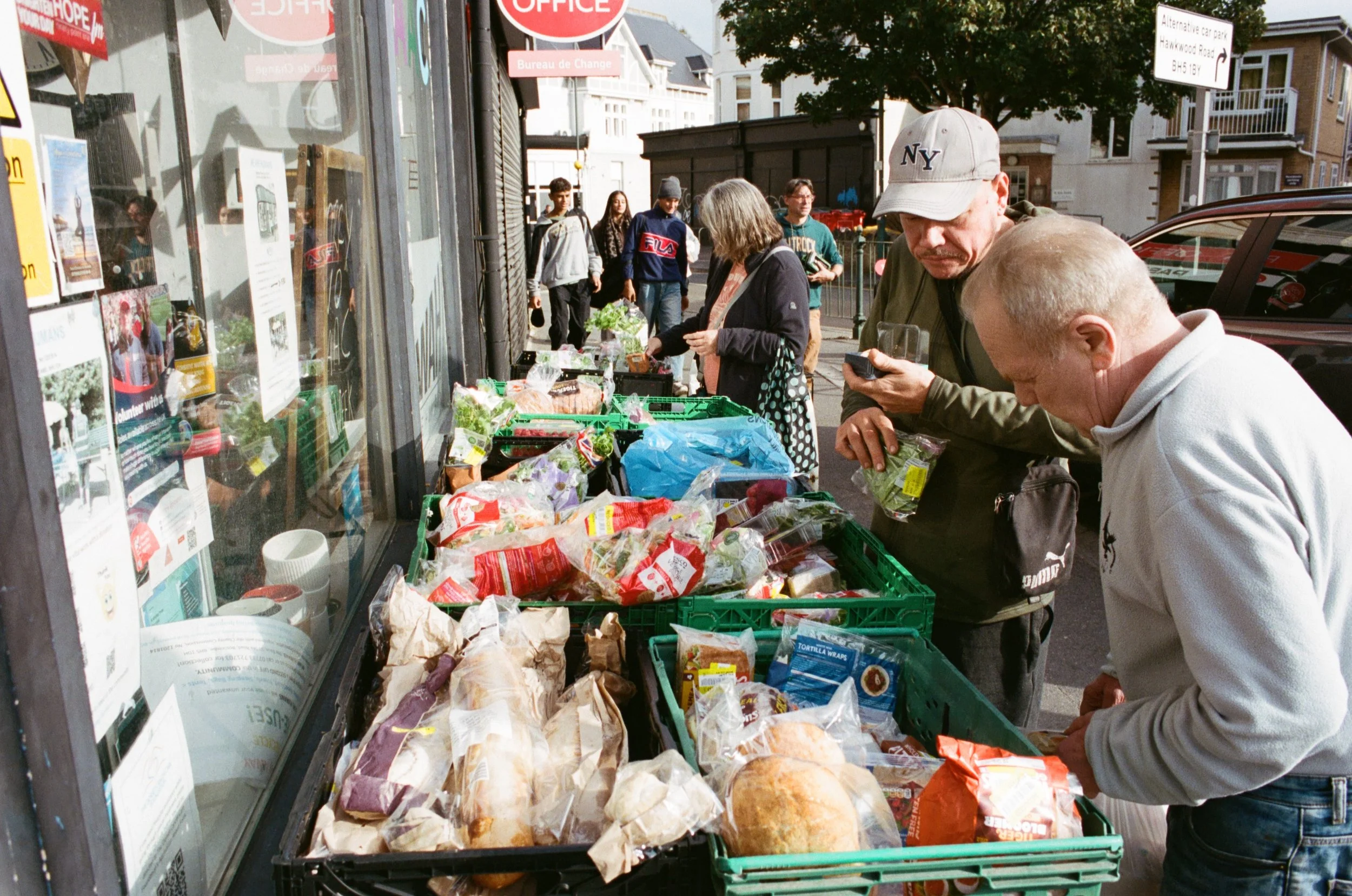 People shopping at a street vendor stand with various snacks and groceries, including bread, chips, and produce, on a sunny day.