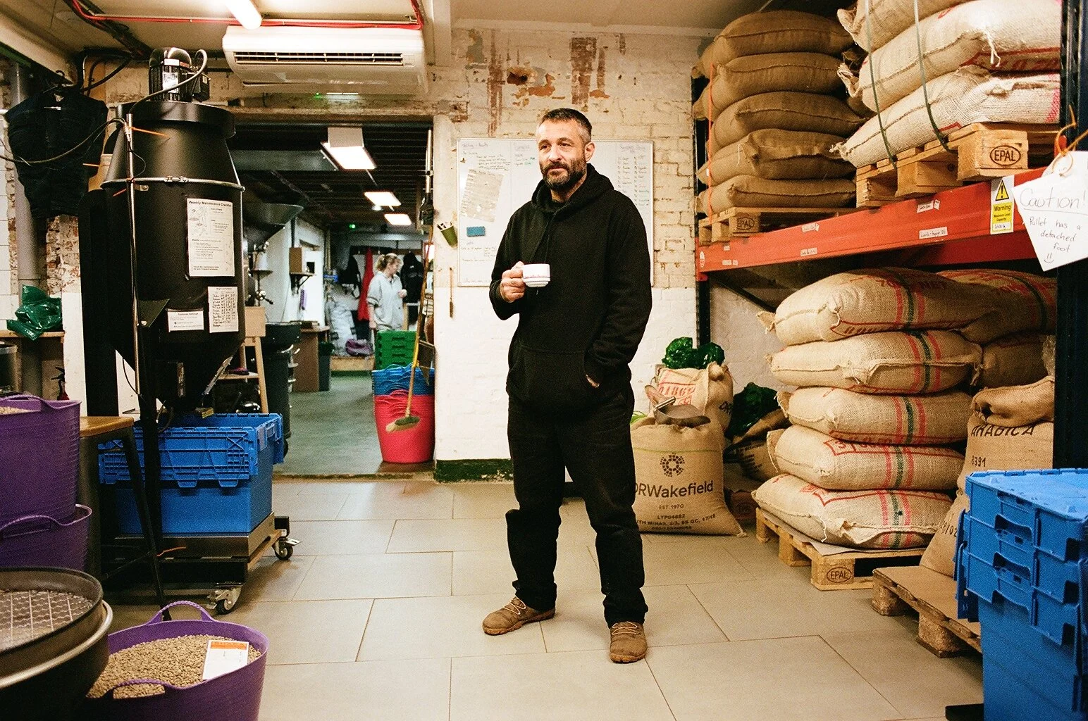Bad Hand Coffee team member holding a coffee cup beside roasting equipment and stacked sacks of coffee beans. Photo by Shahab Notghi.