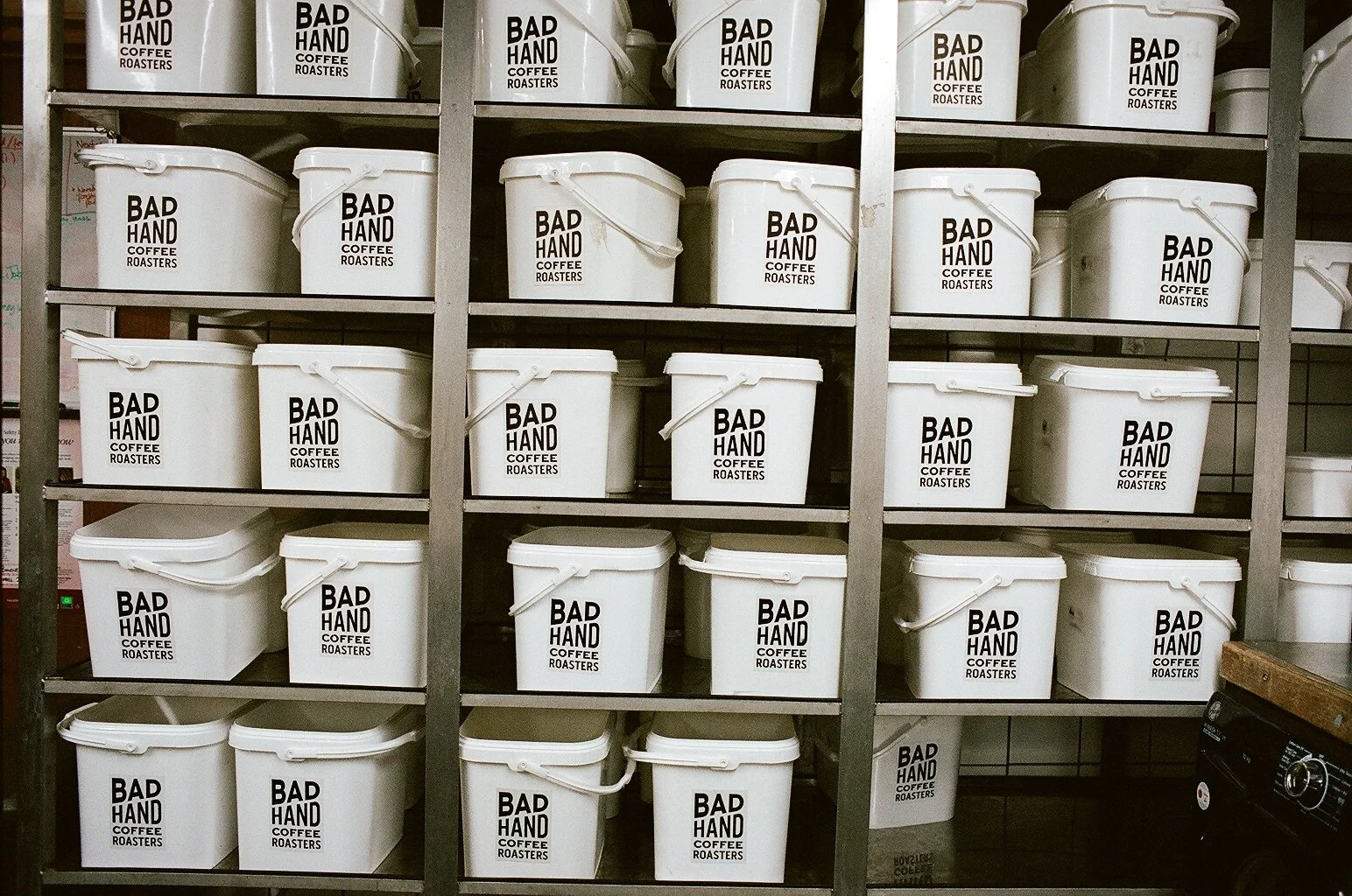 Rows of white storage buckets displaying the Bad Hand Coffee Roasters logo inside the roastery. Photo by Shahab Notghi.
